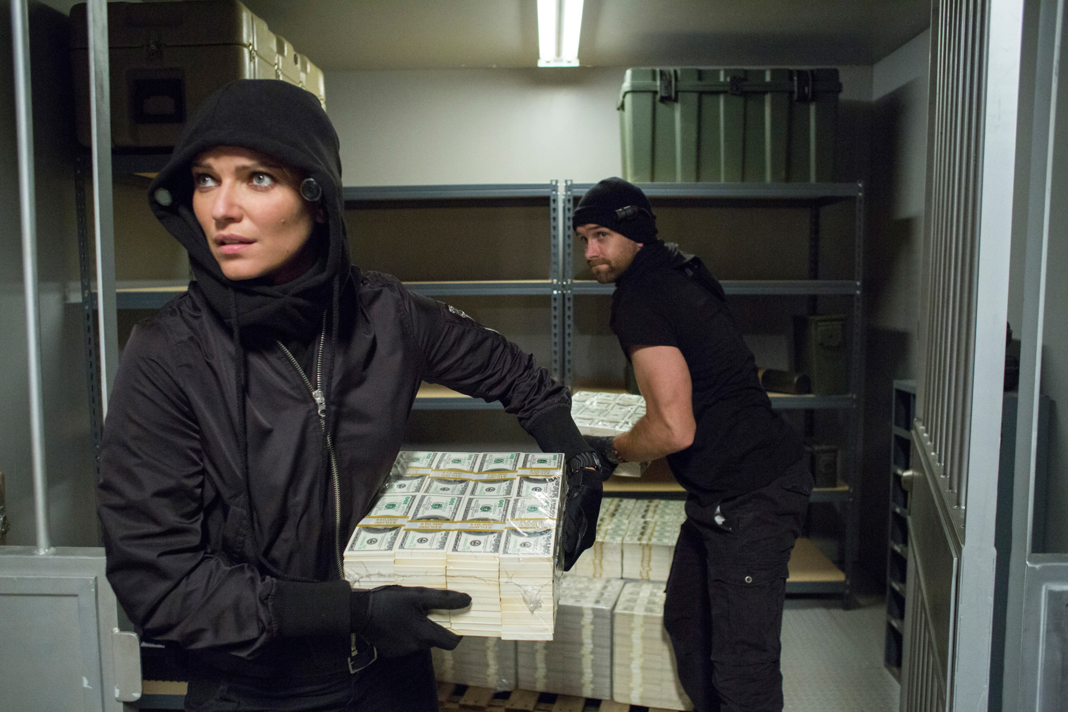Two unidentified individuals dressed in black clothing and ski masks carrying stacks of money in a storage room