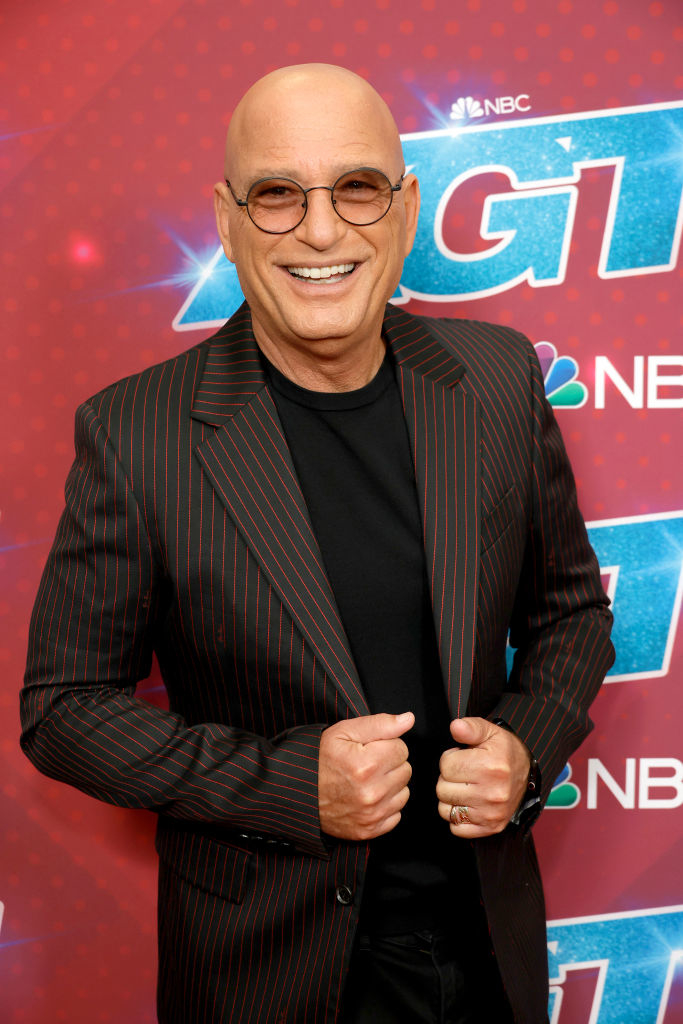Howie Mandel smiles, wearing a black shirt and a dark pinstriped blazer, in front of an 'America's Got Talent' backdrop