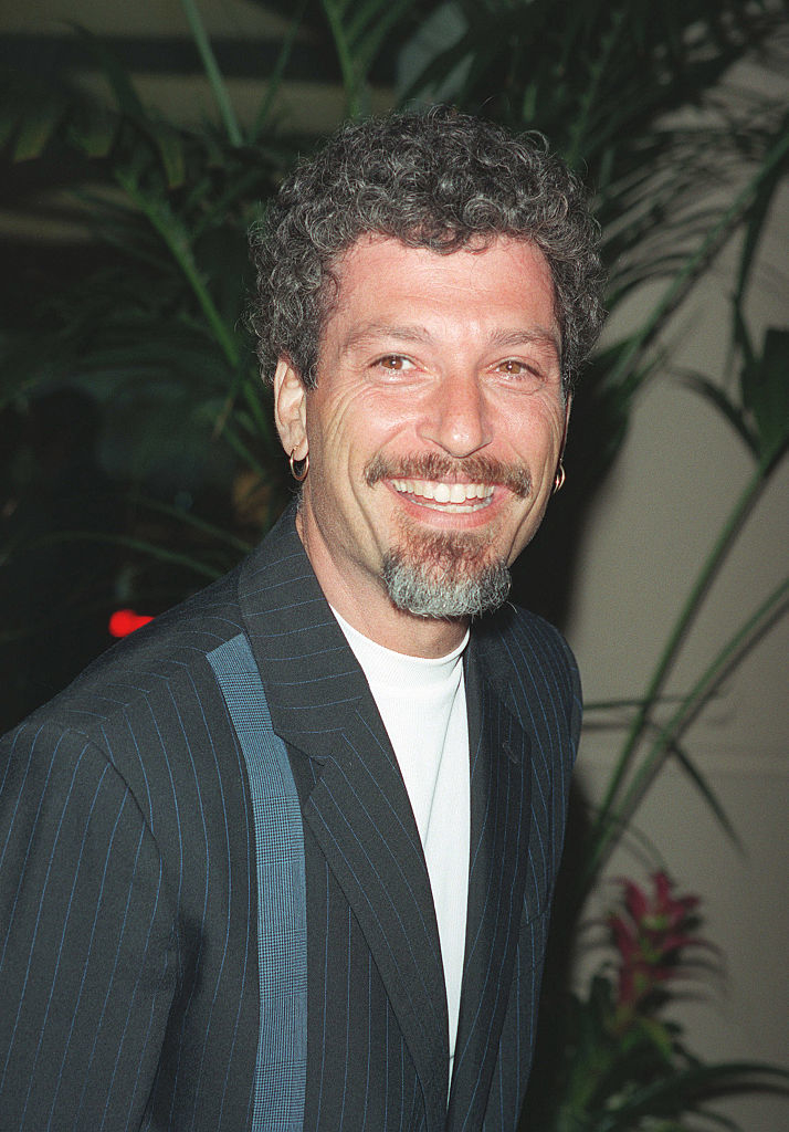 Man with curly hair, goatee, wearing a pinstriped blazer over a white shirt smiles at the camera