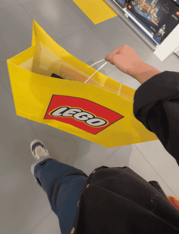 Person holding a large yellow LEGO shopping bag, walking through a store aisle with a visible LEGO display in the background