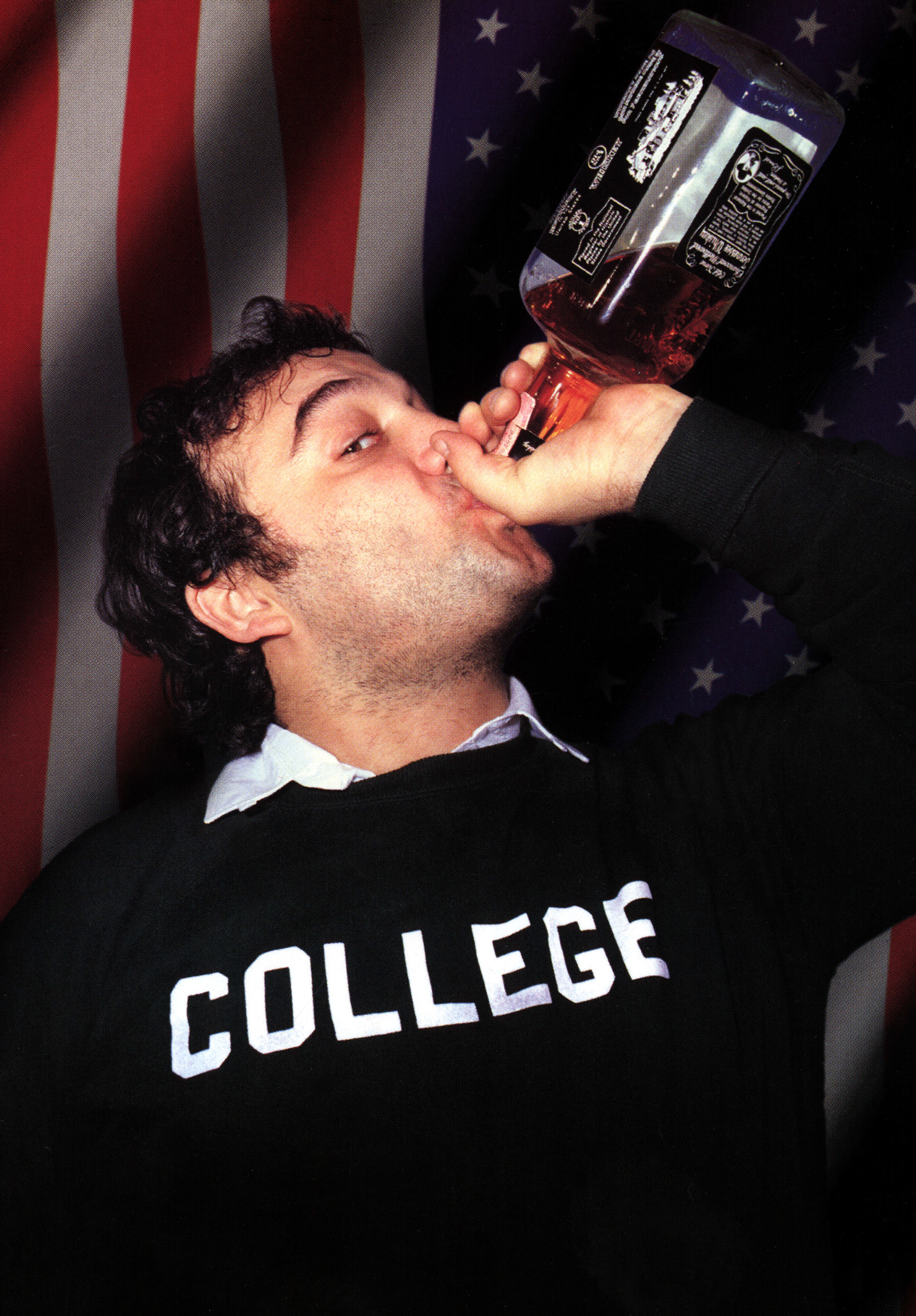 John Belushi in a "College" sweatshirt drinks from a bottle with an American flag backdrop