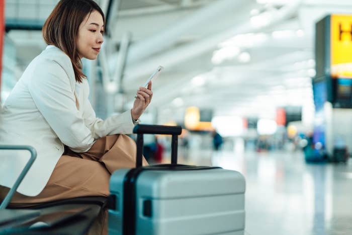 A woman is sitting in an airport terminal next to a suitcase, looking at her smartphone. She is wearing a blazer and is waiting for her flight