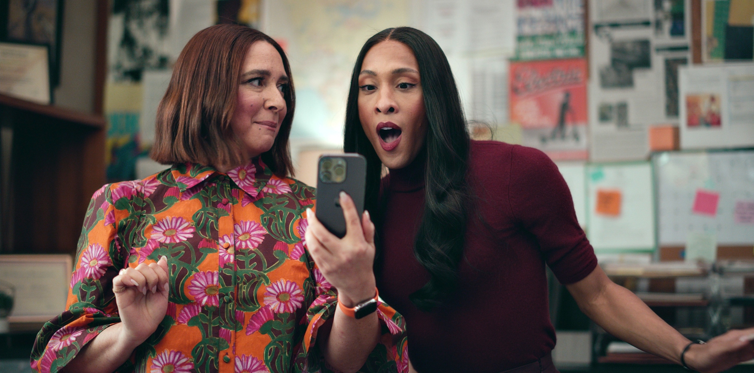 Maya Rudolph and MJ Rodriguez look at a smartphone together, appearing surprised and amused. Maya wears a floral-patterned top, and MJ wears a solid-colored top