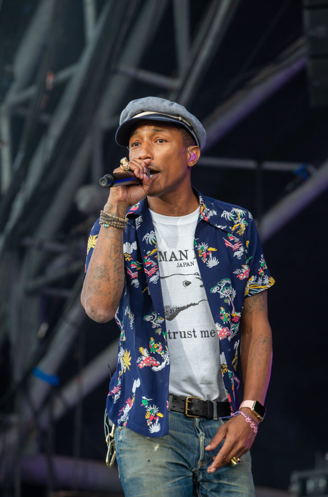 Pharrell Williams performs on stage wearing a short-sleeve printed shirt, white t-shirt, jeans, and a grey cap while holding a microphone