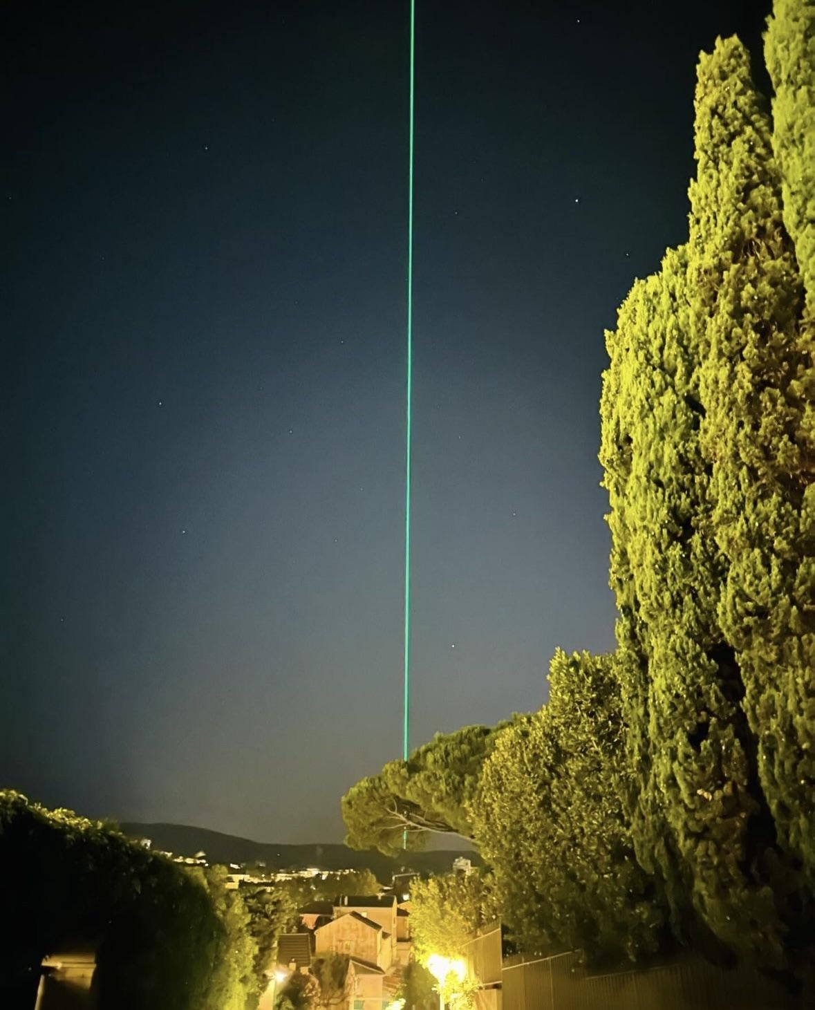 A bright green laser beam stretches vertically into the night sky, illuminating a serene residential area surrounded by tall trees