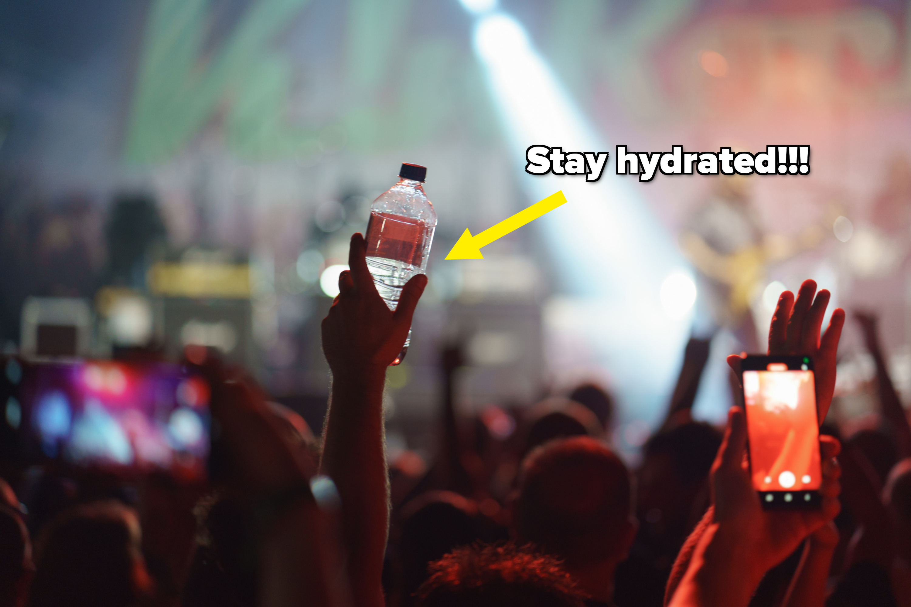 Concert audience with hands raised, one holding a water bottle and another taking a photo with a smartphone. Stage and performers blurred in the background