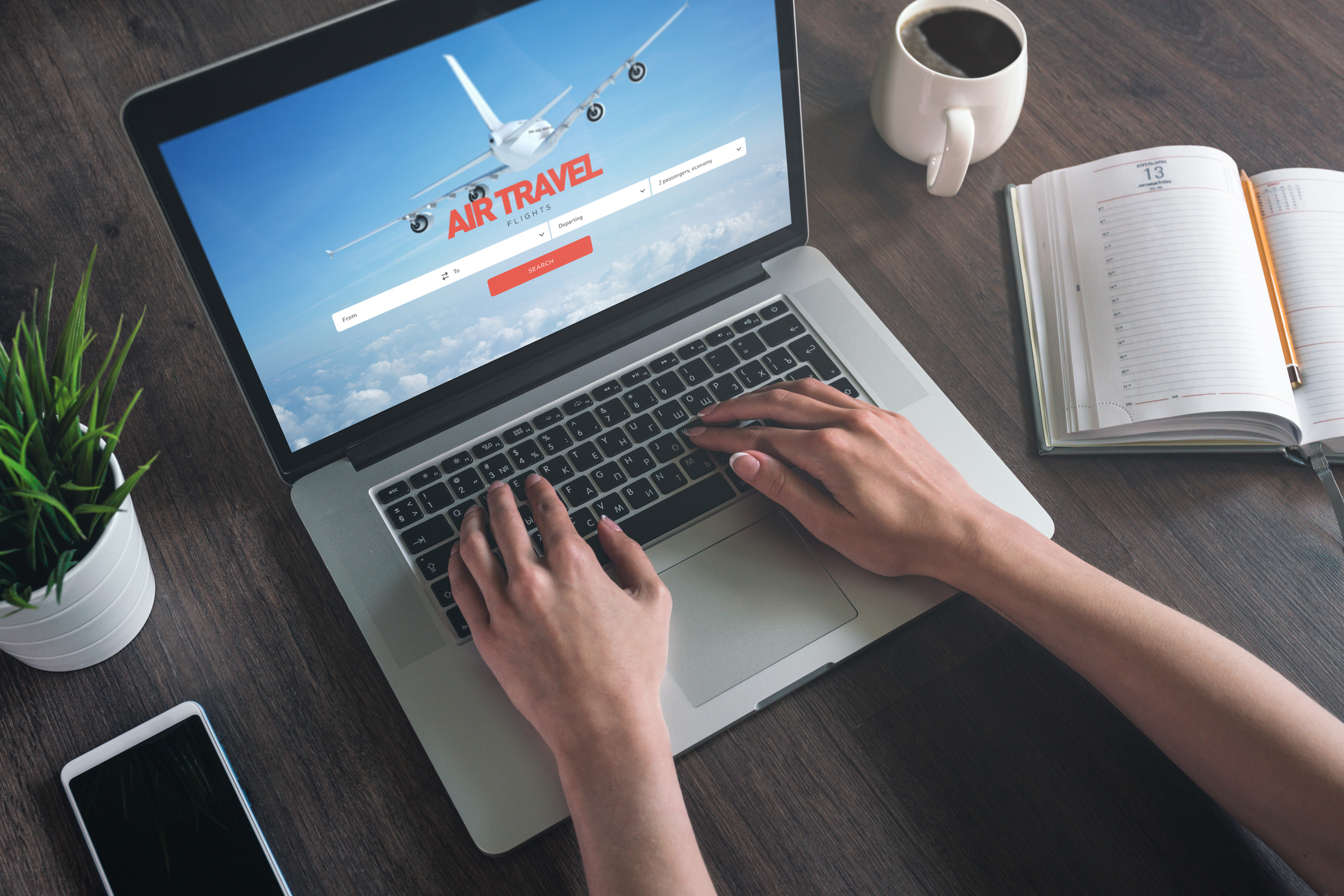 A person is using a laptop with an air travel booking website open. The table holds a phone, plant, planner, and cup of coffee