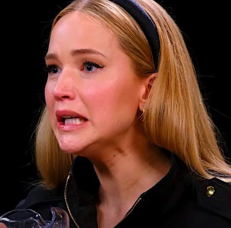 Jennifer Lawrence appears distressed while holding a glass of water, wearing a black outfit with a headband
