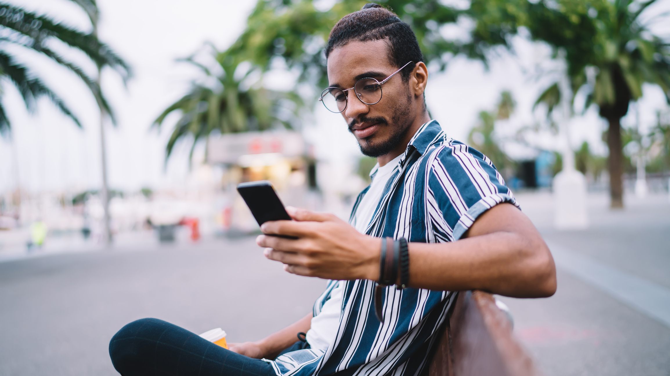 Person at a tropical destination sitting on a bench, looking at their phone, holding a coffee. Palm trees and a blurred building can be seen in the background