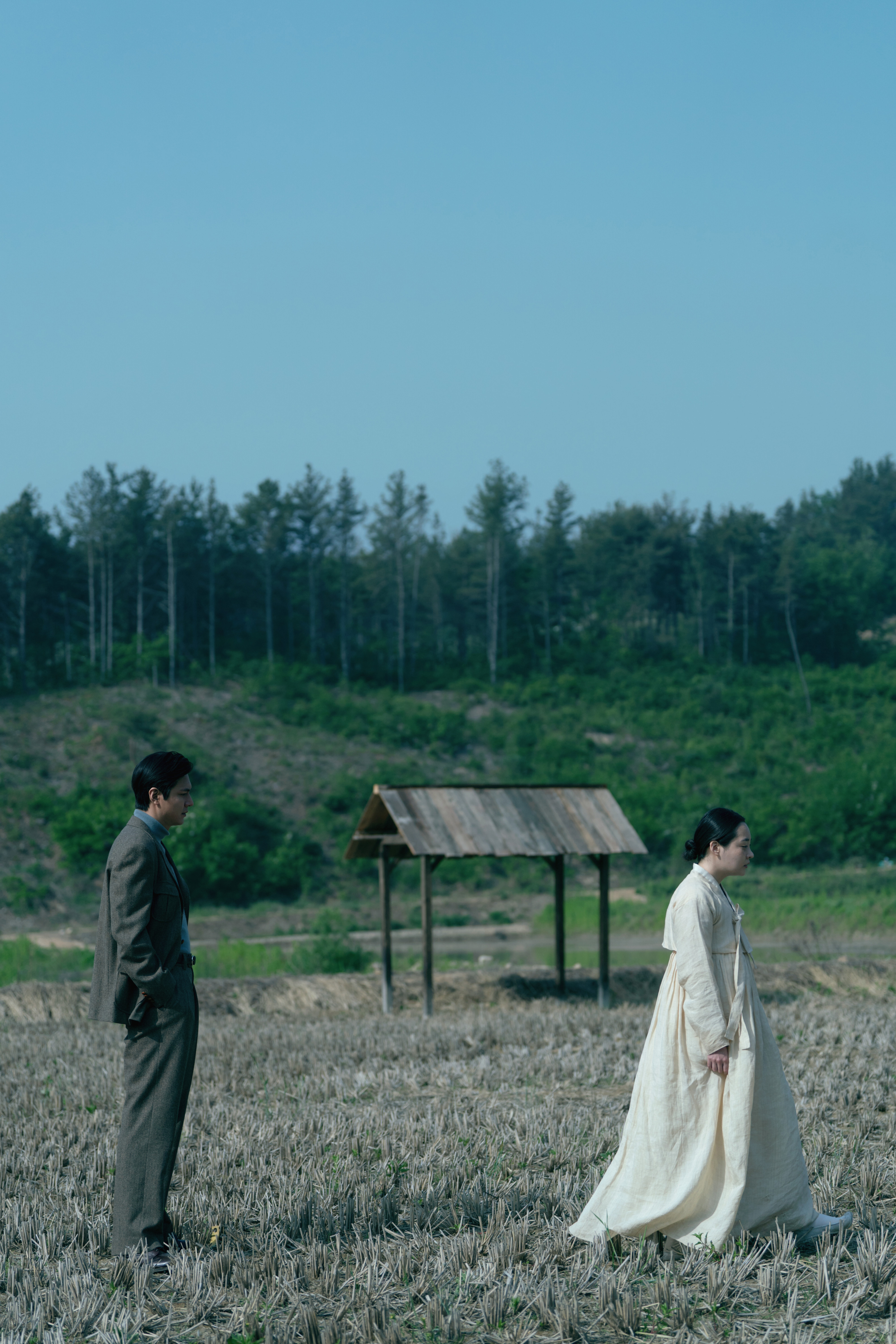 Lee Min-ho and Minha Kim in traditional attire, standing in a rural field with a small wooden structure in the background