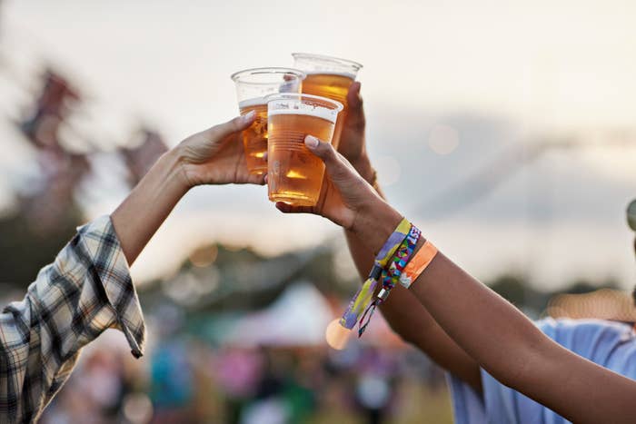 Hands holding cups of beer for a toast at an outdoor festival, with wristbands visible. The image captures a celebratory moment in a lively setting
