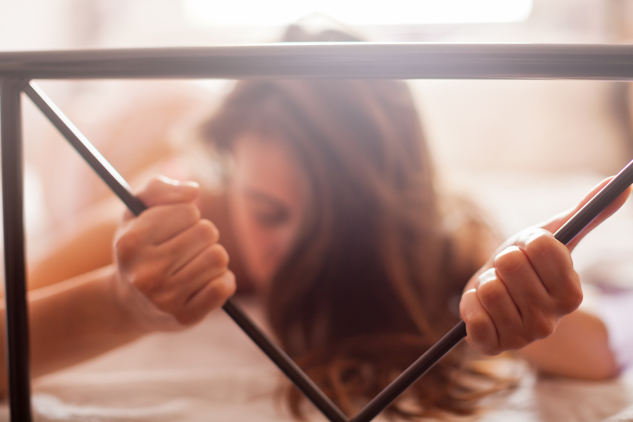 A person gripping a bed frame with both hands, head lowered and long hair falling forward, suggesting an intimate or emotional moment