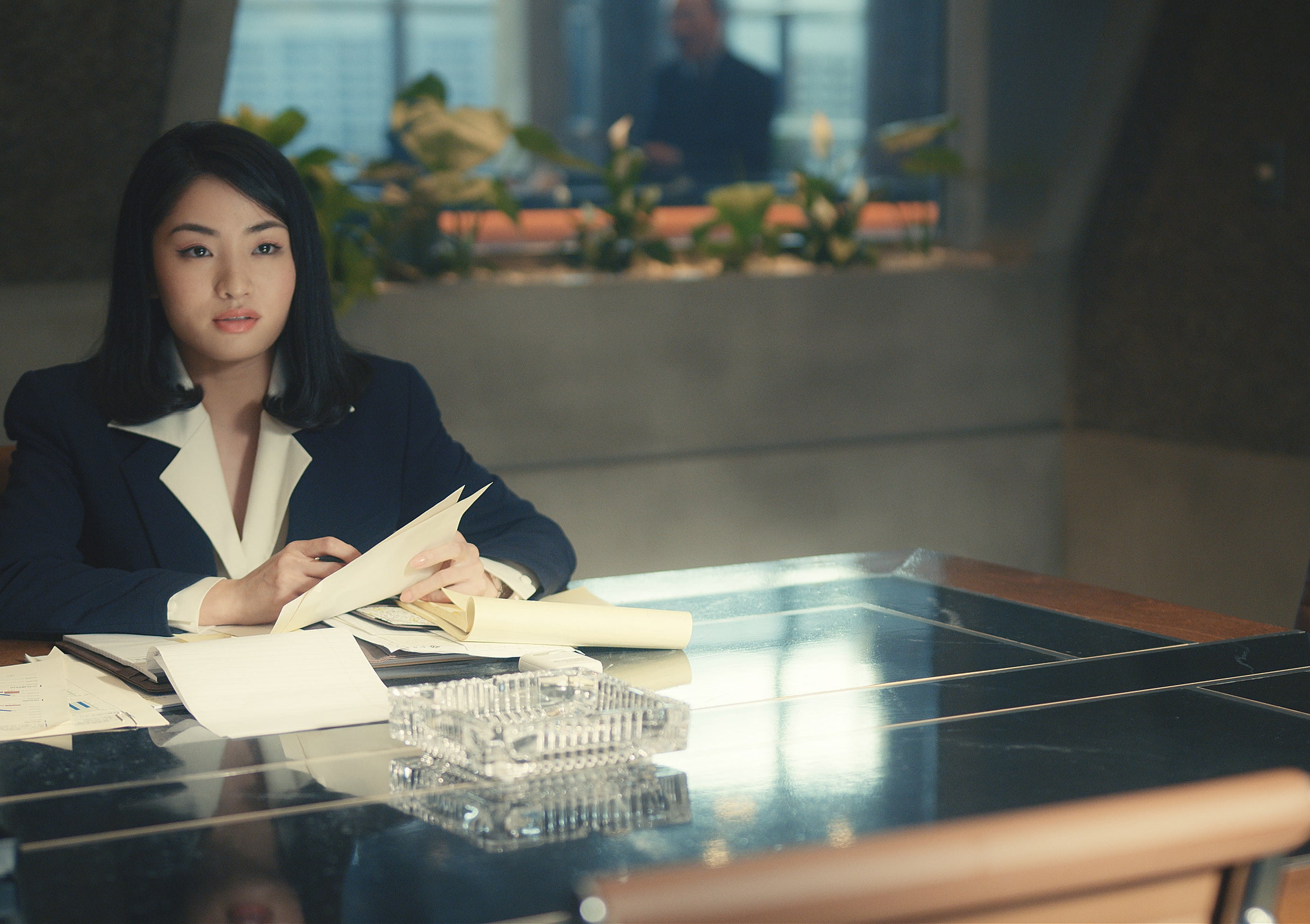 Anna Sawai in "Pachinko" seated at a desk with documents in a modern office setting, looking focused