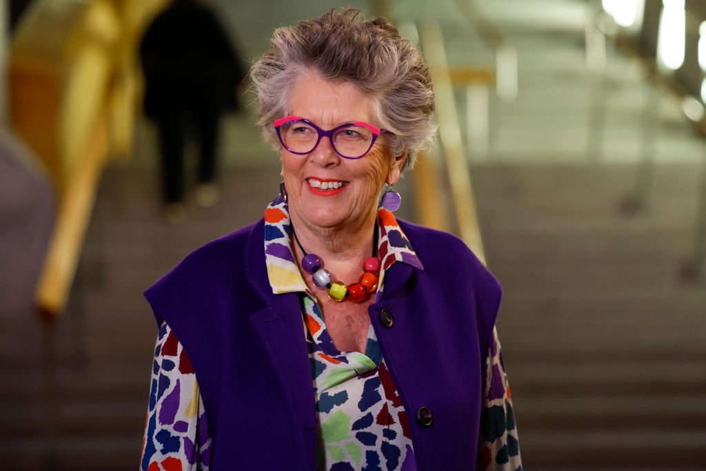 Prue Leith smiling on a staircase, wearing colorful, patterned clothing with a chunky necklace and purple glasses