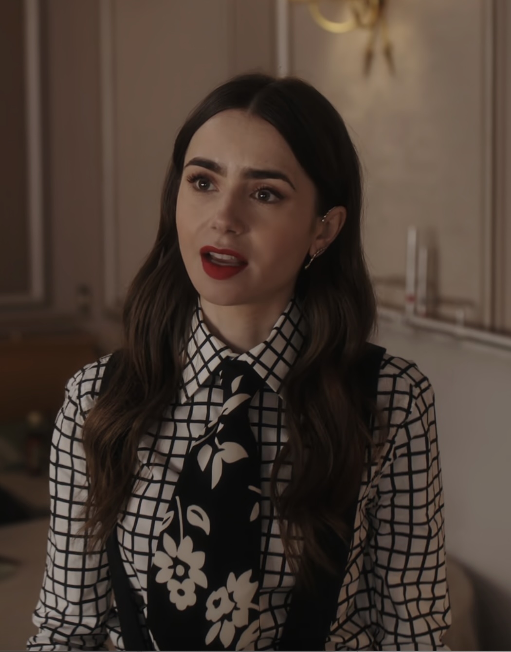 Lily Collins wearing a checkered blouse with a large floral tie, appears to be talking
