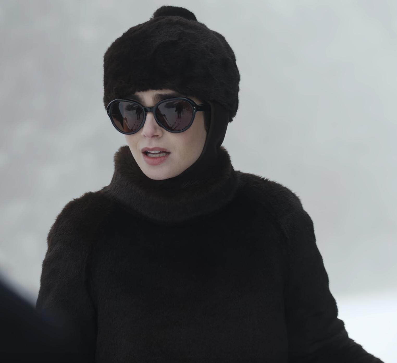 Lily Collins wearing a fur hat, sunglasses, and a turtleneck, set against a snowy background.