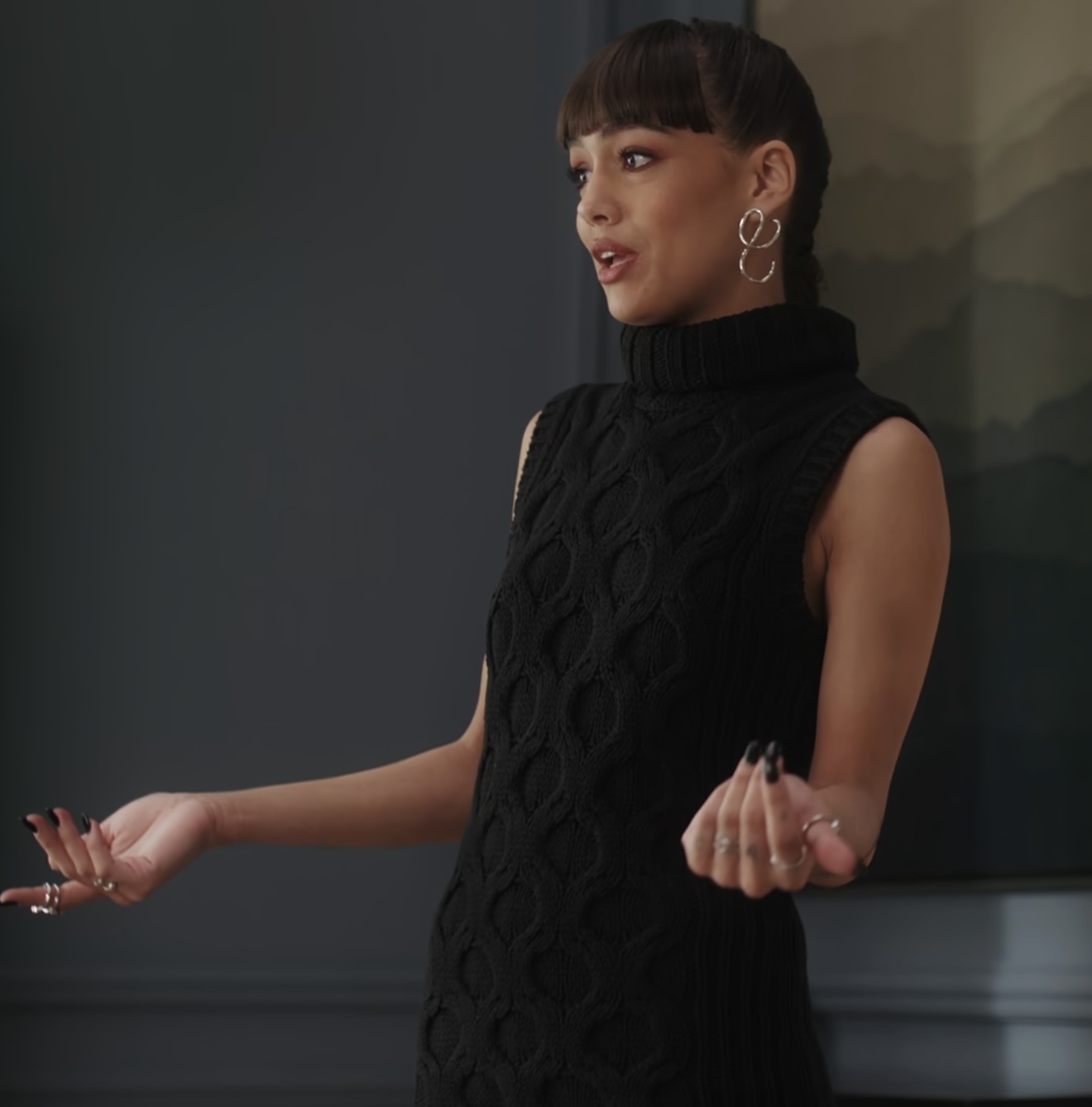 A person stands indoors, gesturing with their hands, and wearing a sleeveless textured turtleneck sweater with large hoop earrings. Name unknown