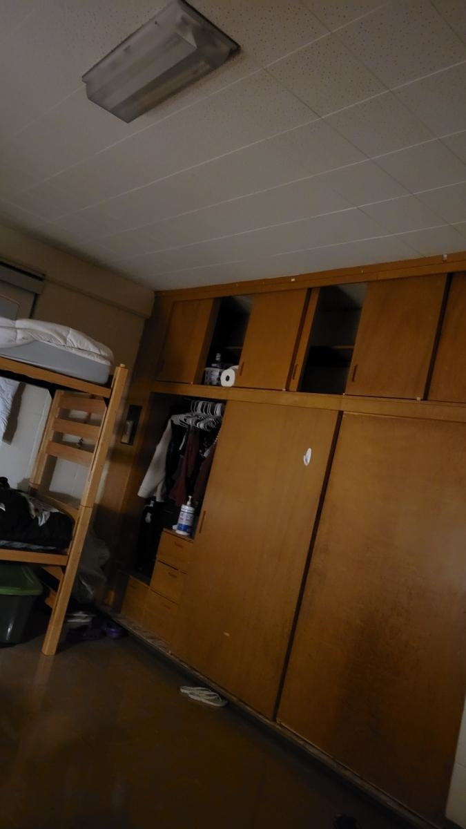A cluttered dorm room with a bunk bed on the left, an open closet in the center, and wooden cabinets overhead. Various clothing items are visible