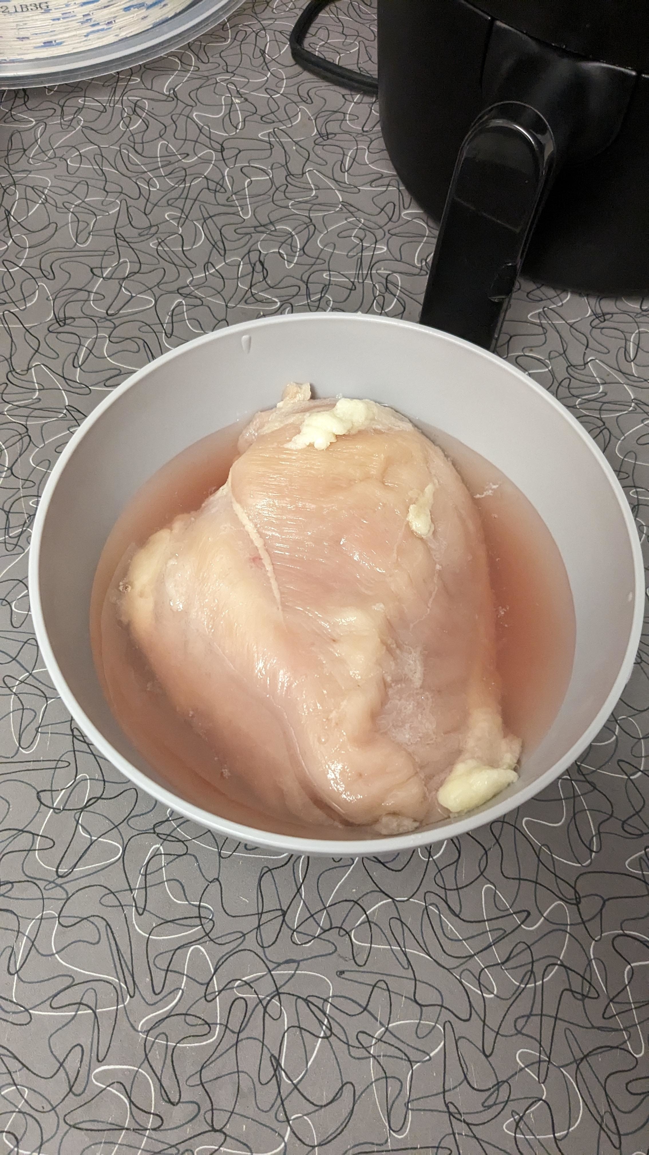 Raw chicken breast soaking in a bowl of water. A black kitchen appliance is placed on the countertop in the background
