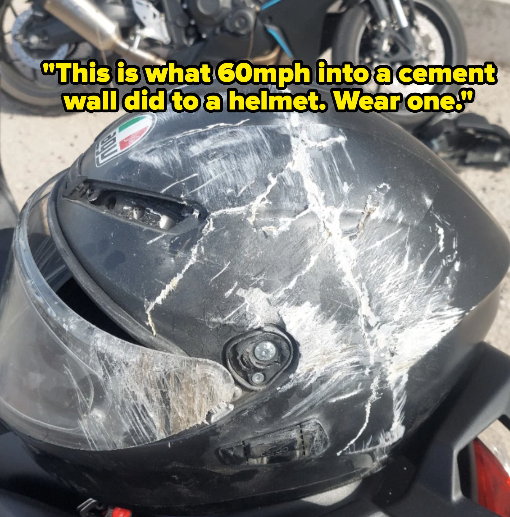 A scratched motorcycle helmet, showing significant damage. Two motorcycles are parked in the background