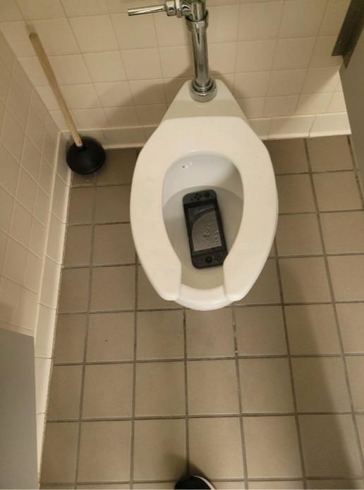 A toilet with a mobile phone at the bottom of the bowl and a plunger nearby on the tiled floor