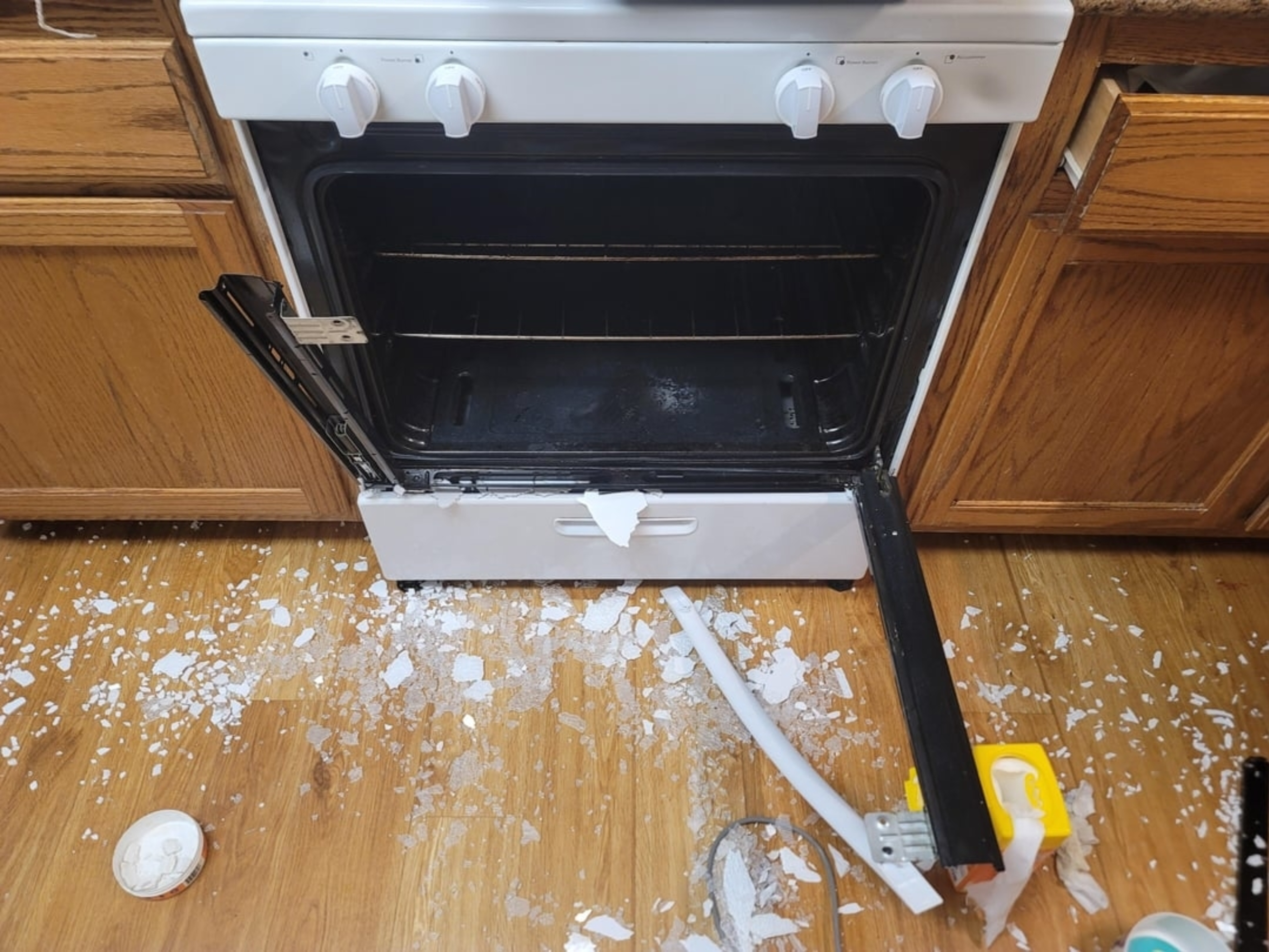 A kitchen oven with a broken glass door, shattered glass on the floor, and nearby kitchen cabinets partially visible