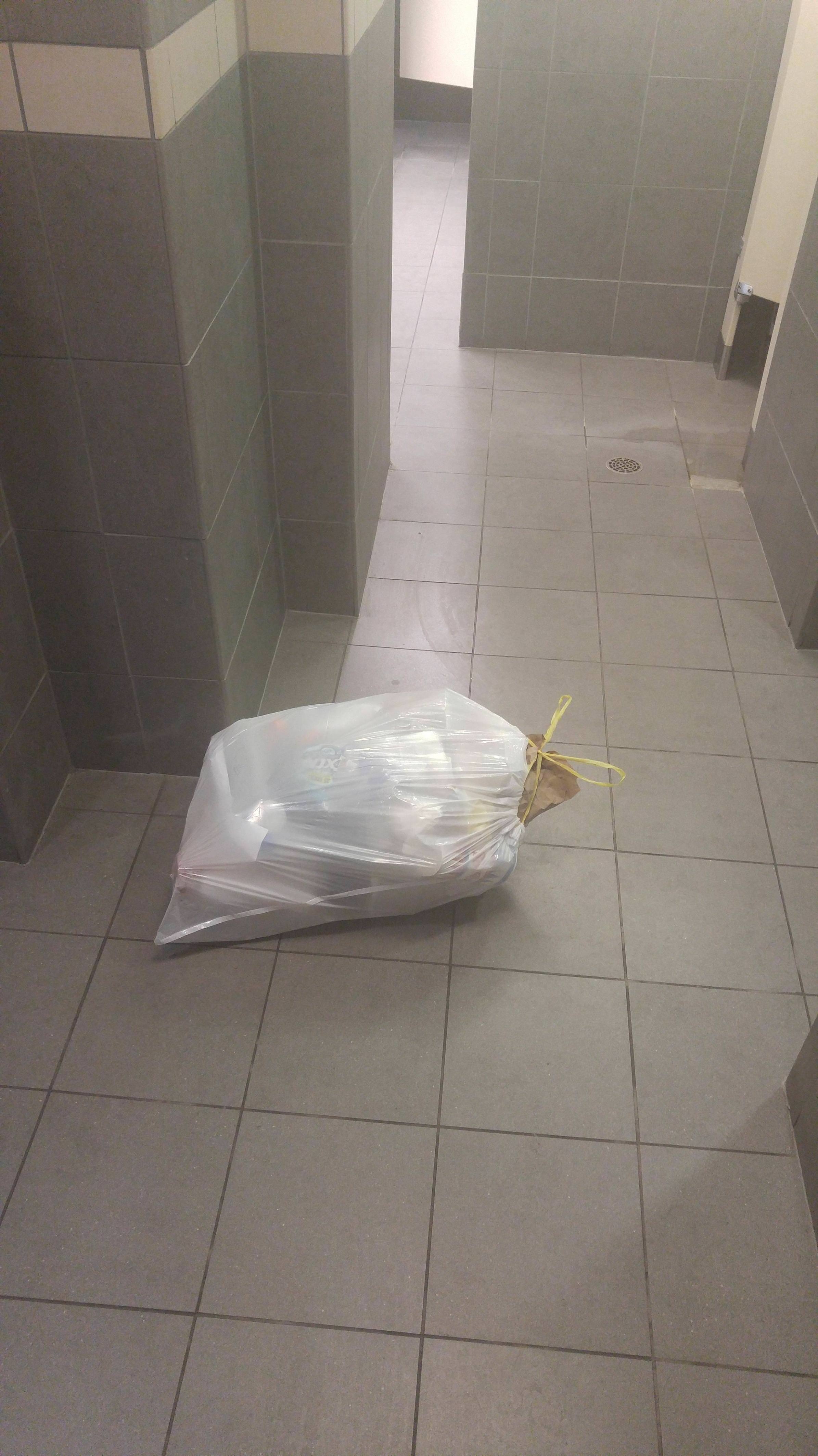 A bag of trash is left on the floor of a tiled public restroom. The bag appears full and is tied at the top