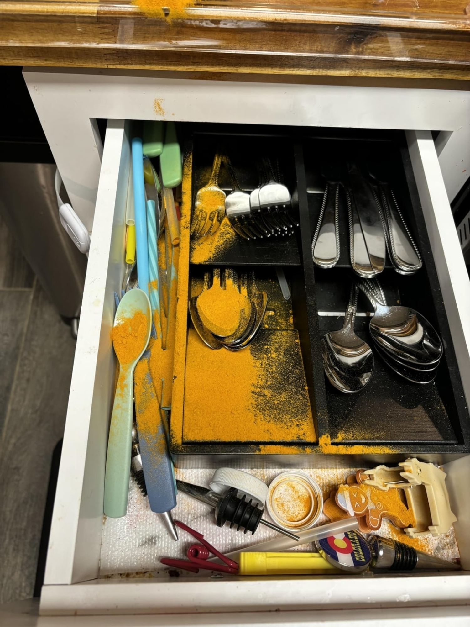 Open kitchen drawer with silverware and various utensils covered in spilled turmeric powder