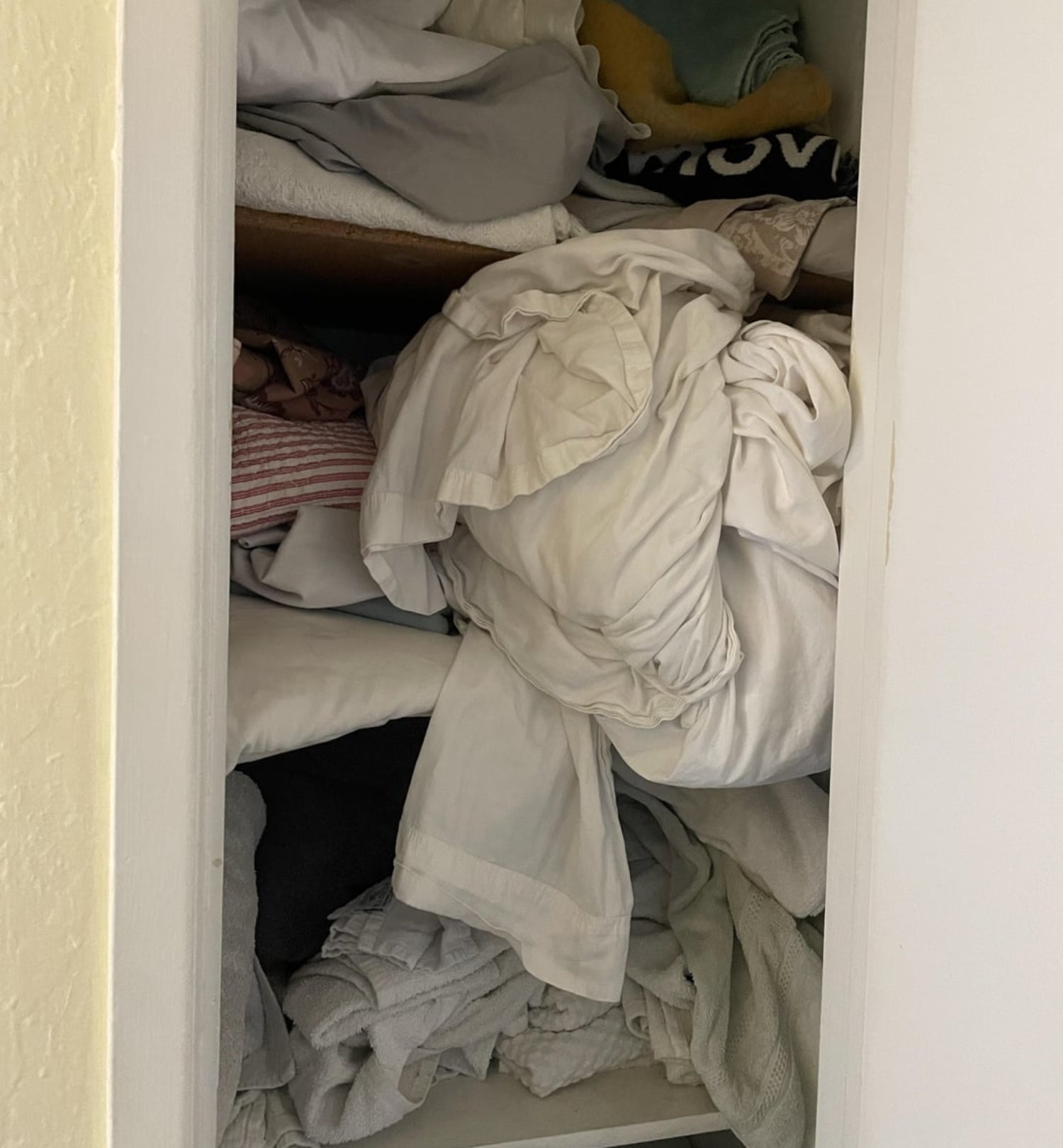 Linen closet filled with piles of disorganized towels, blankets, and sheets, and a fitted sheet falling off a shelf