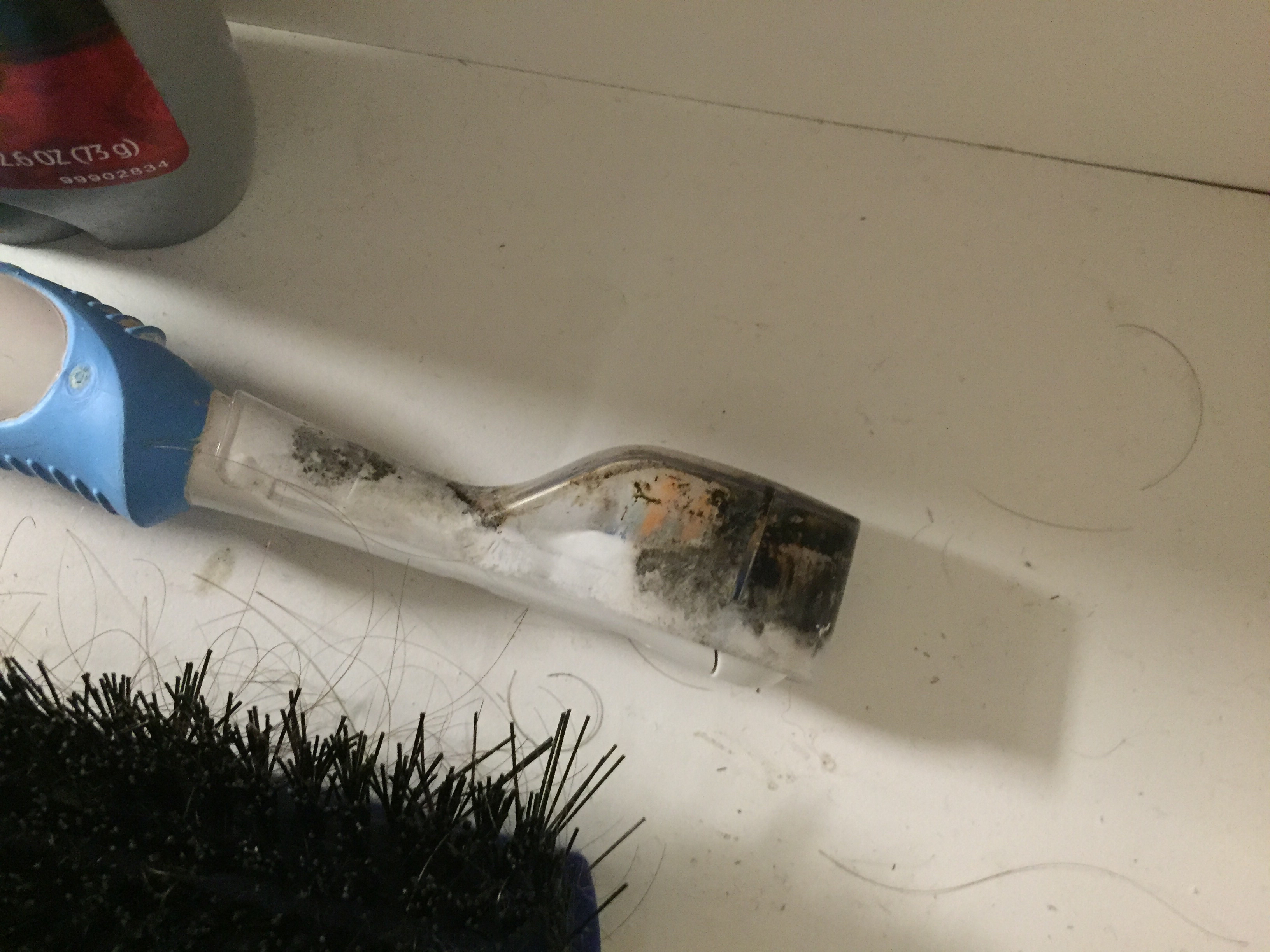 Electric toothbrush head is covered in mold and grime, lying on a white surface next to a black-bristled brush and a bottle of cleaning product