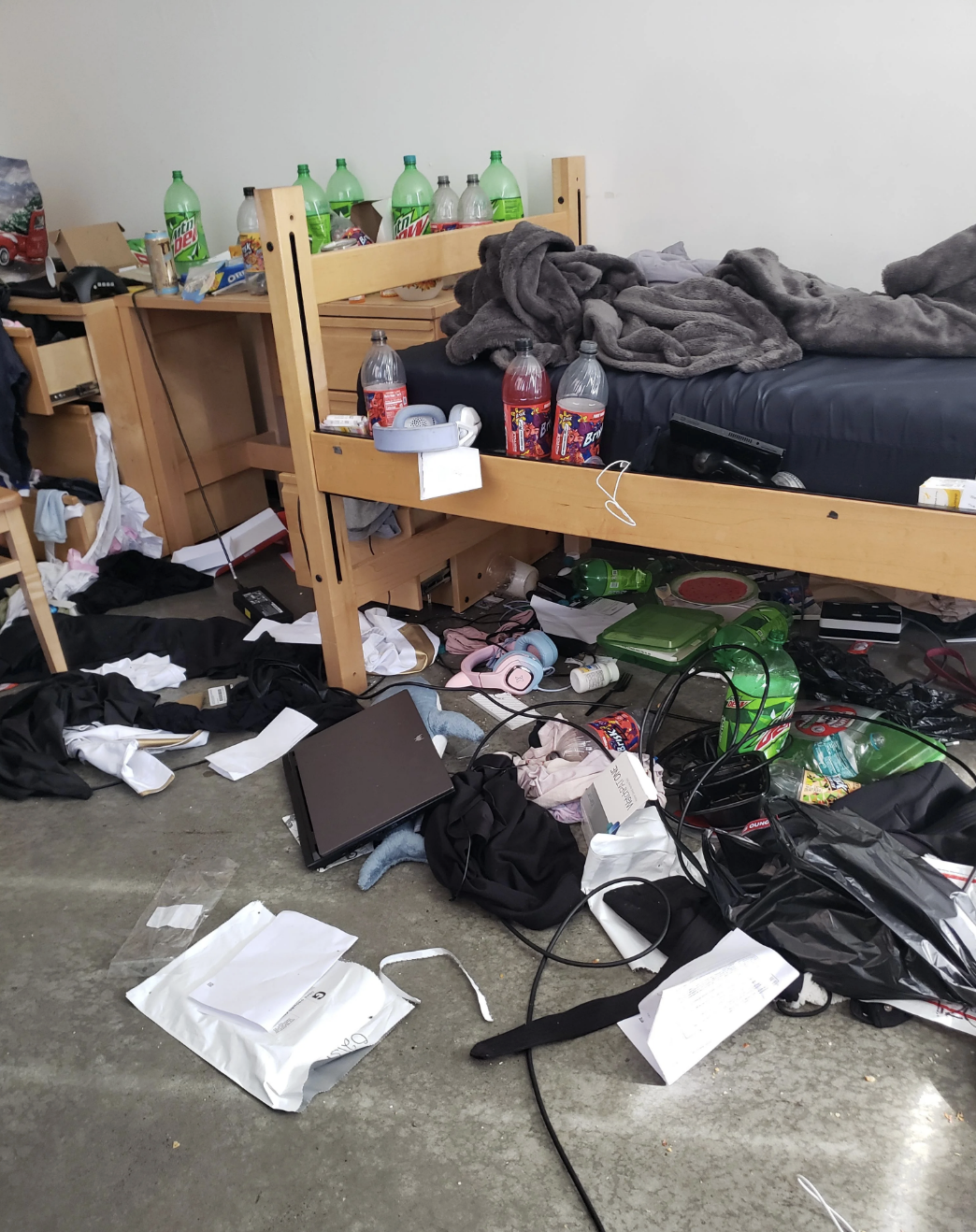 Messy dorm room with items scattered everywhere, including bottles, trash, clothes, electronics, and a cluttered desk next to a bunk bed