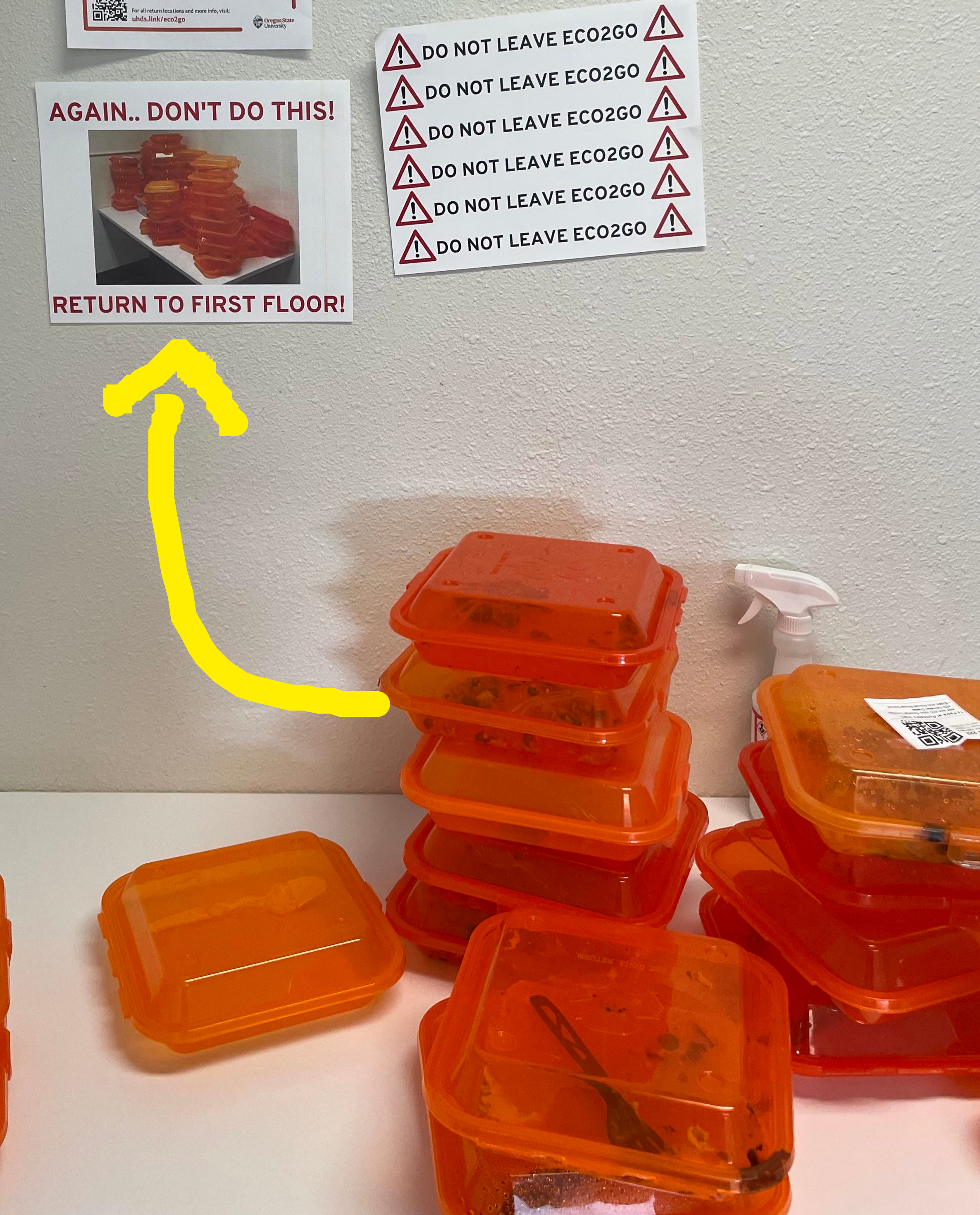 Stack of orange ECO2GO containers on a counter with notices urging not to leave containers and to return them to the first floor