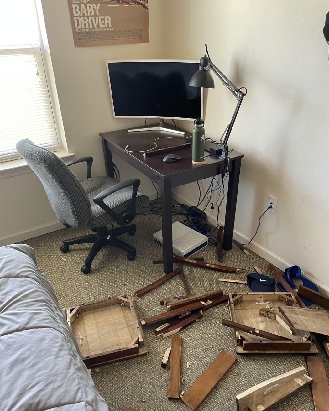Broken pieces of a wooden table are scattered on the floor next to a desk with a computer monitor, a lamp, a water bottle, and an office chair