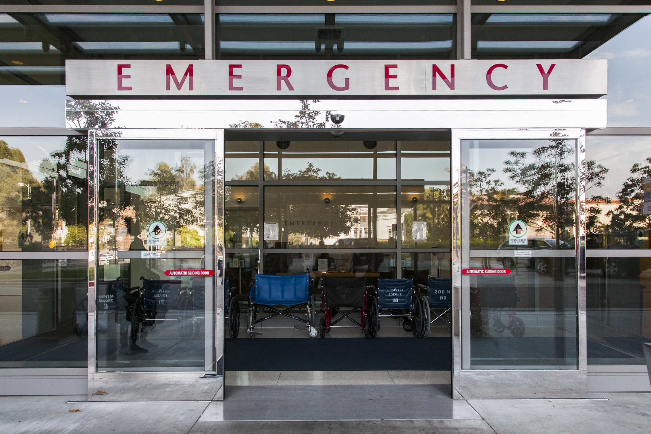 Emergency entrance with glass doors and signage in a medical facility. Visible inside are wheelchairs near the reception area