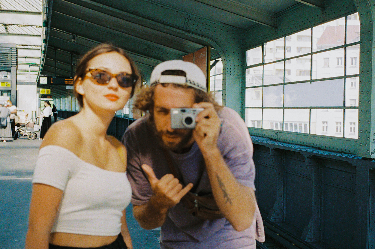 Two people on a train platform; the woman in a white off-shoulder top, sunglasses, and the man in a backwards cap, taking a photo with a camera