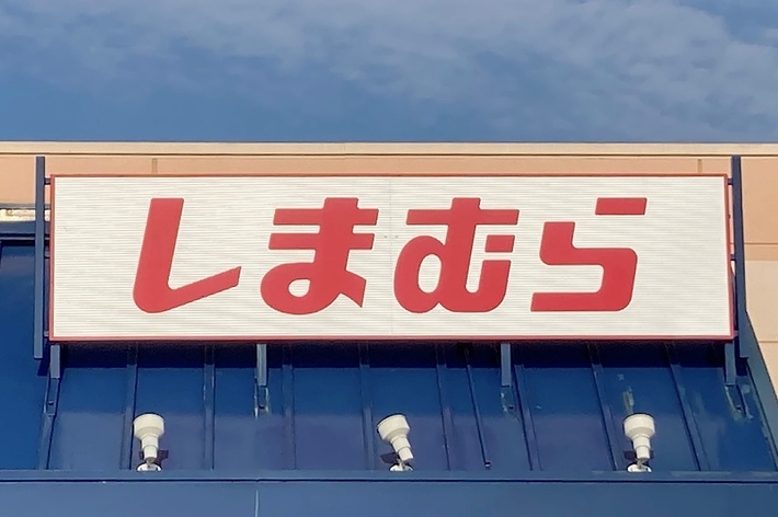 The image shows a store sign with Japanese text, reading &ldquo;しまむら,&rdquo; above the entrance of a building