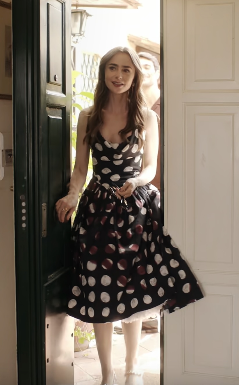 Lily Collins stands smiling in a doorway, wearing a polka dot dress with a fitted bodice and flared skirt