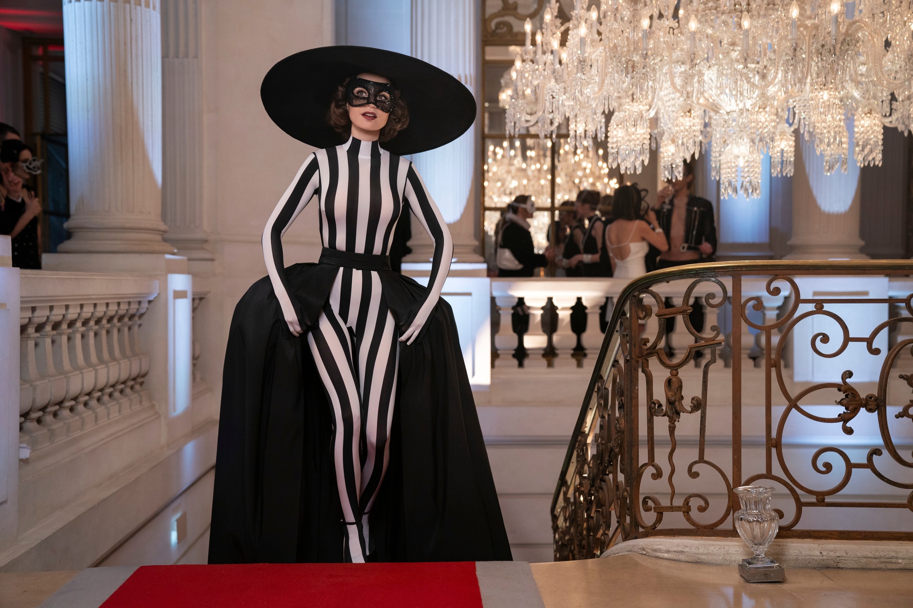 Woman wearing a black and white striped dress, large black hat, and mask standing on a red carpet at an elaborate, chandelier-lit event