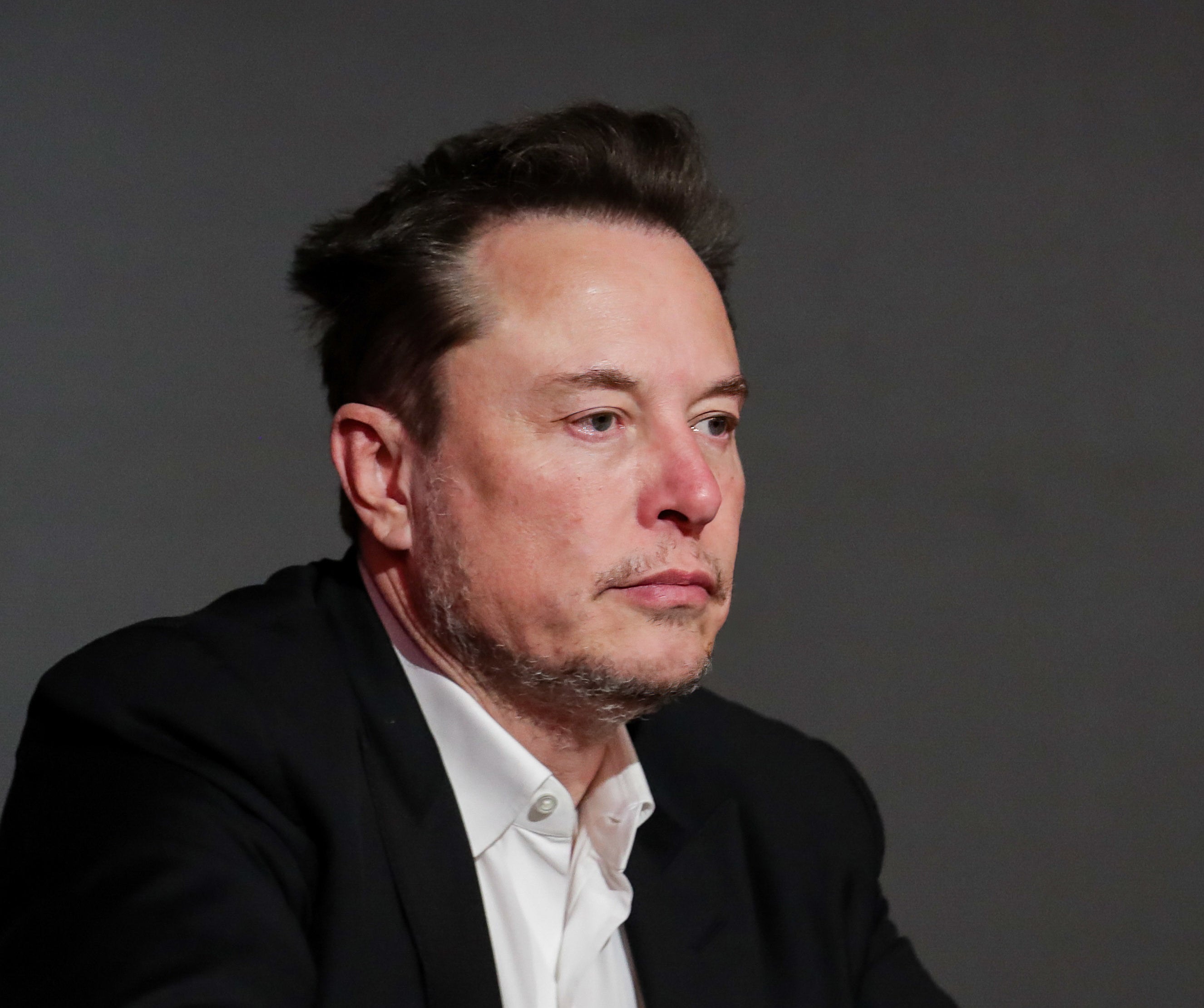 Elon Musk in a dark suit jacket and white shirt, looking thoughtful