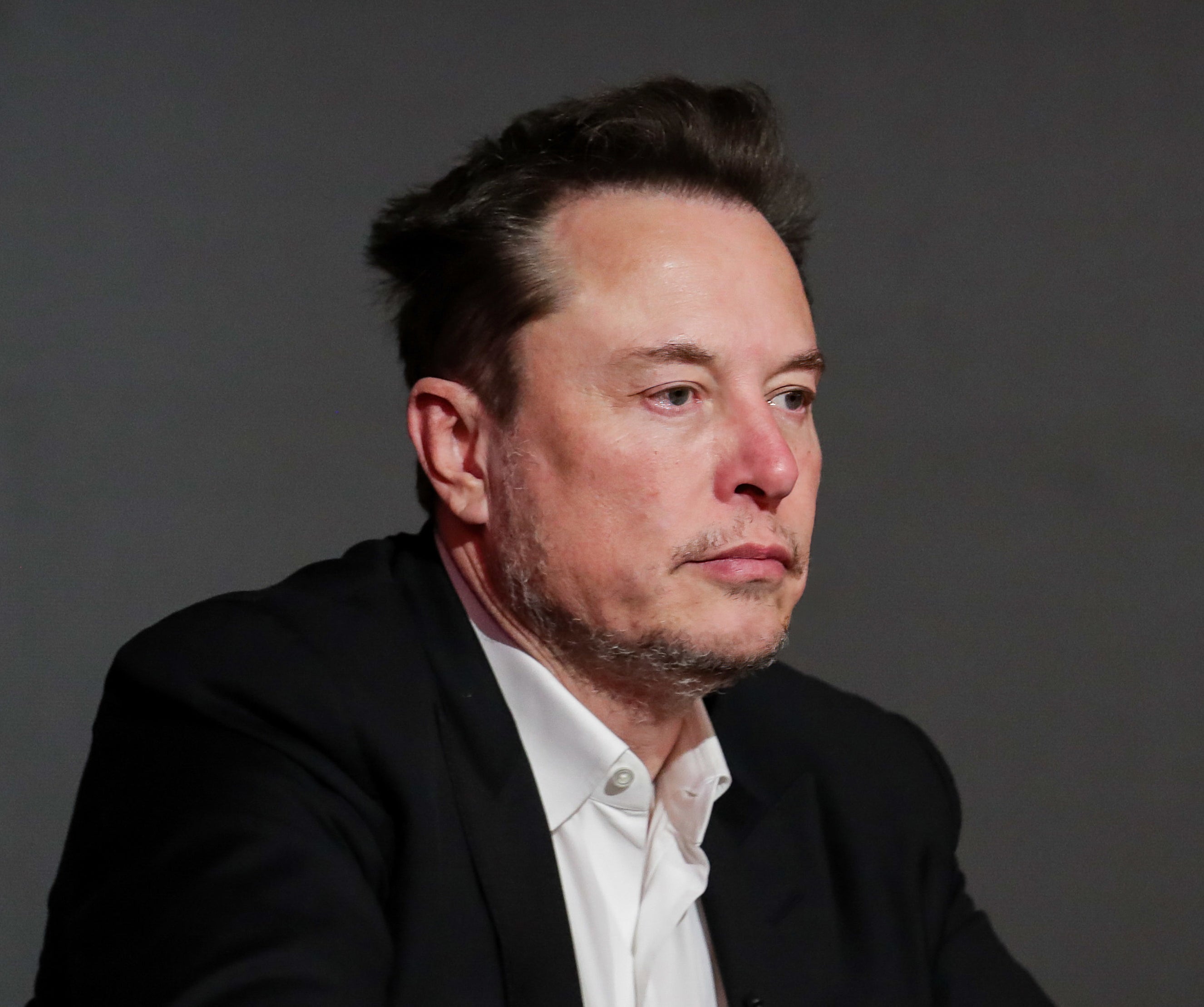 Elon Musk in a dark suit jacket and white shirt, looking thoughtful