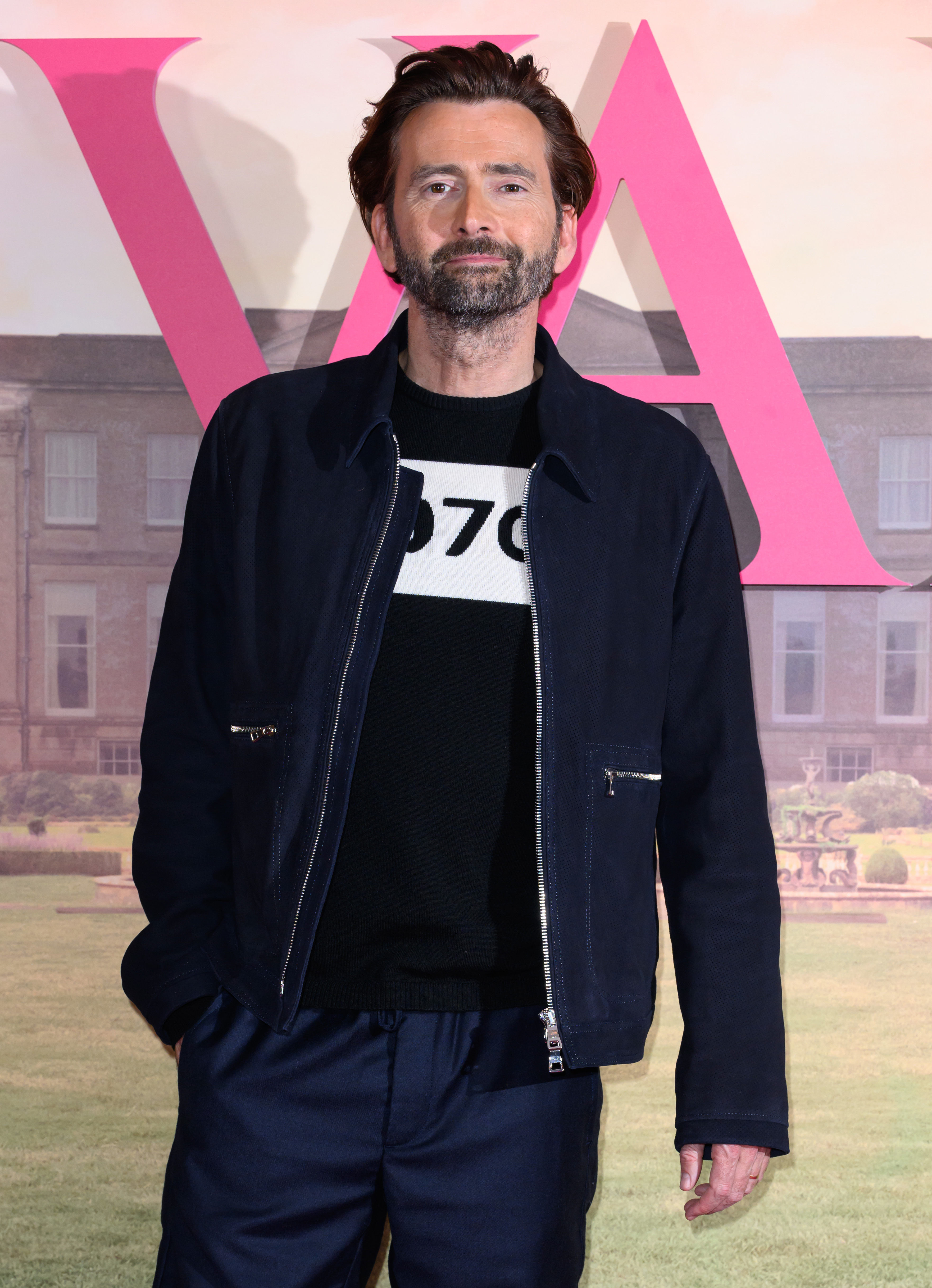 David Tennant in casual attire with hands in pockets, standing in front of a backdrop featuring the letters "VA" and a scenic landscape