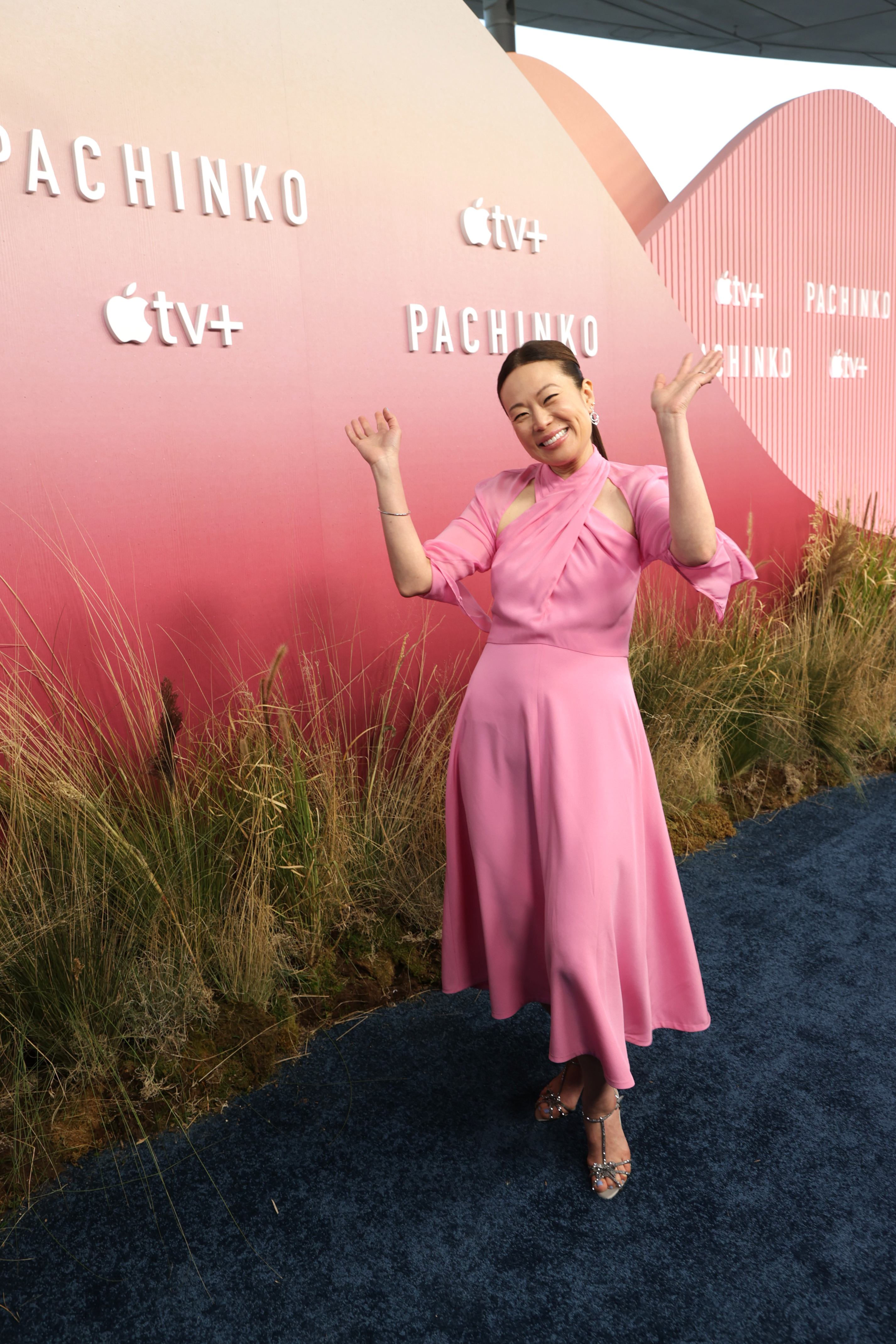 Soo Hugh on the red carpet for the "Pachinko" premiere, wearing a pink dress, smiling, and posing with raised hands