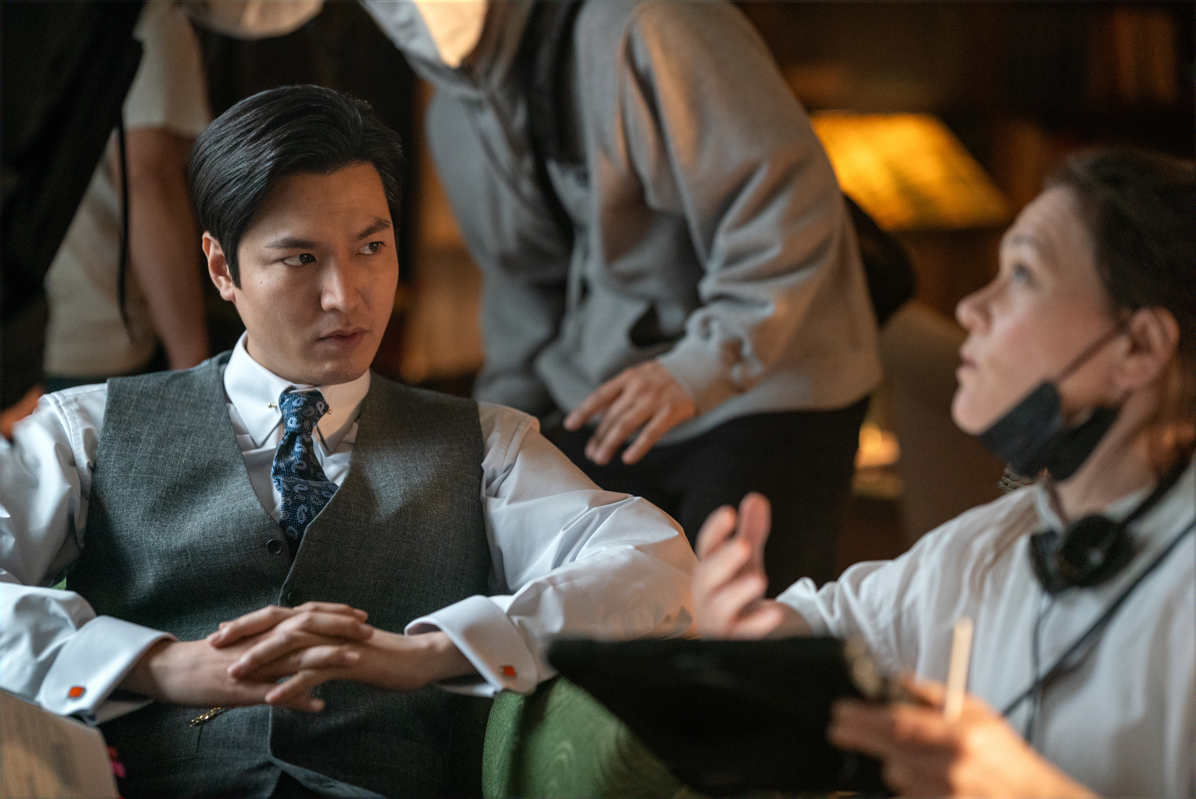 Lee Min-ho, wearing a suit vest and tie, is seated and engaged in conversation with a production crew member on a TV or movie set
