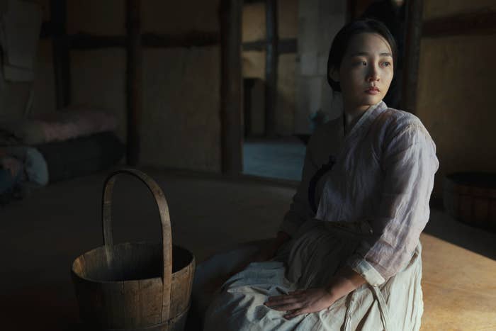 Sunja, portrayed by Kim Min-ha, dressed in traditional Korean clothing, sits indoors next to a wooden basket, looking contemplative