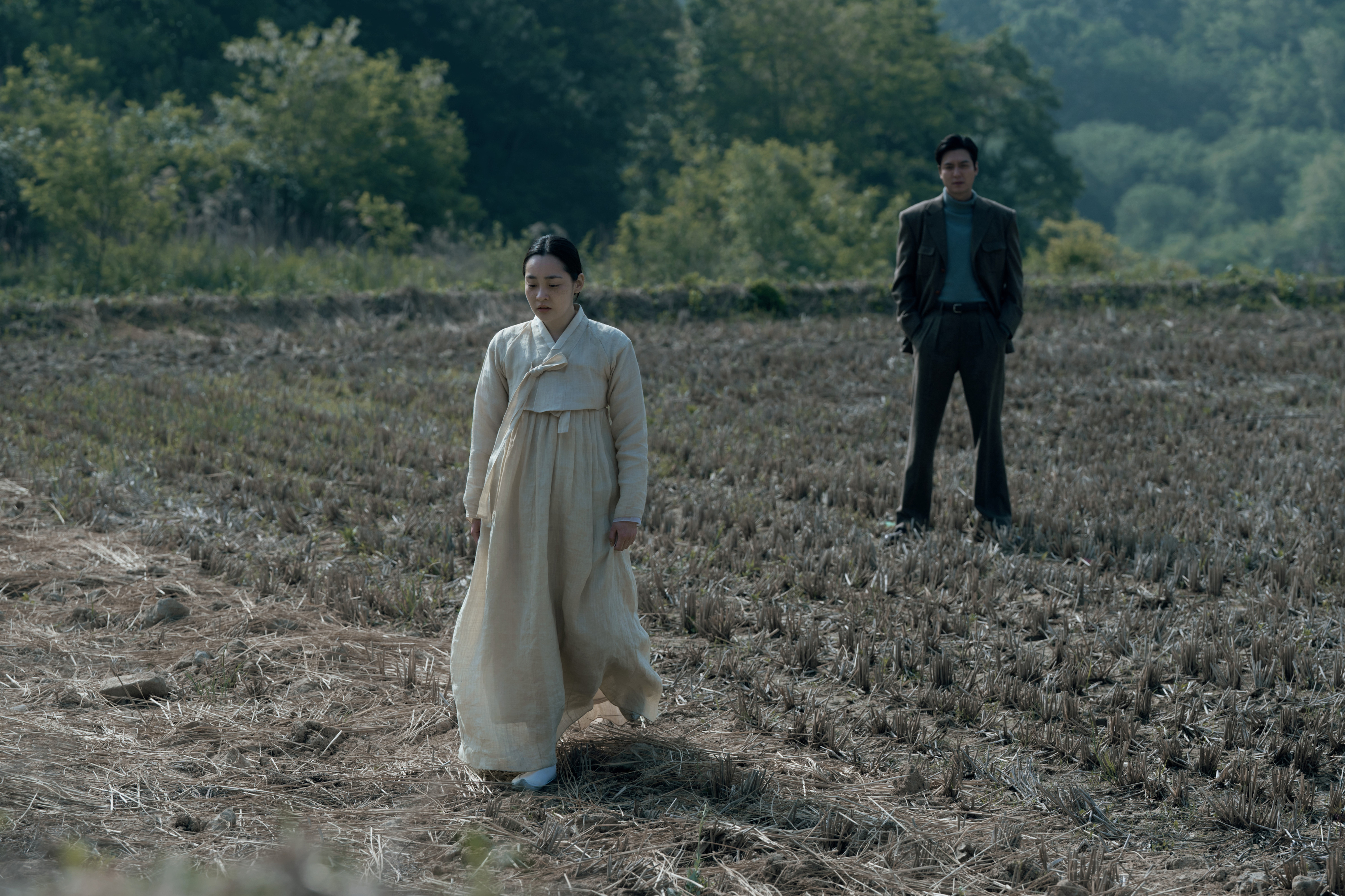 Scene from "Pachinko": Sunja, in traditional attire, walks ahead in a field with Hansu, in a suit, standing in the background, greenery behind them