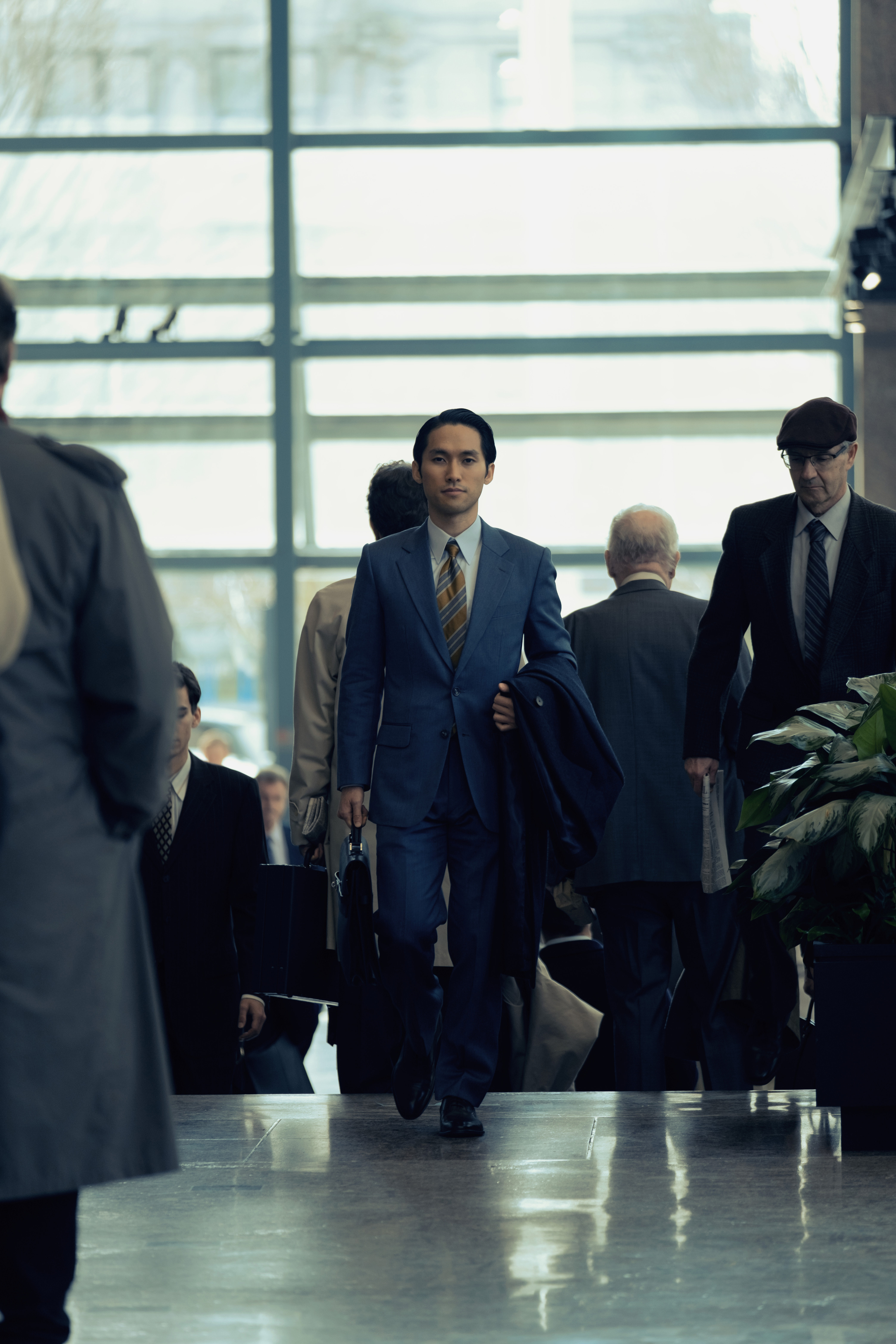 Jin Ha walks purposefully through a lobby in a tailored suit holding a coat and briefcase, amidst a crowd of people. Scene from "Pachinko"