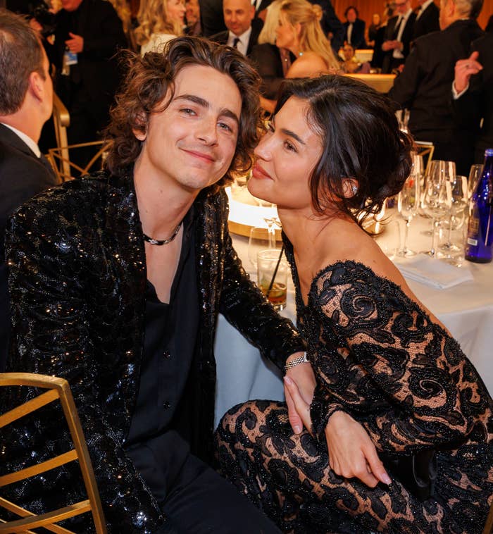 Timothée Chalamet in a black sequined jacket and Sarah Paulson in a lace dress, seated and smiling at an elegant event with others in the background