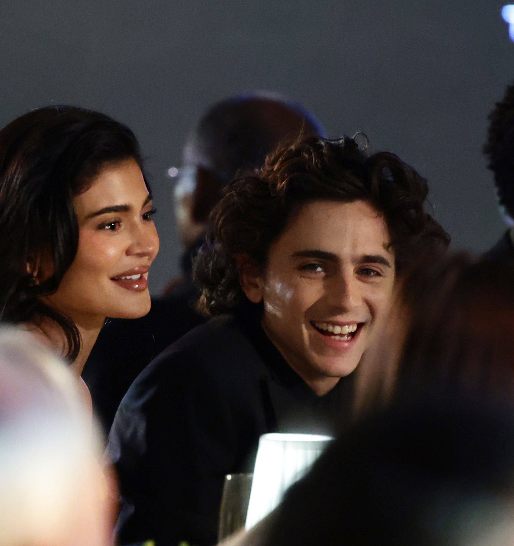 Kylie Jenner and Timothée Chalamet are smiling and sitting together at an event with several other people in the background