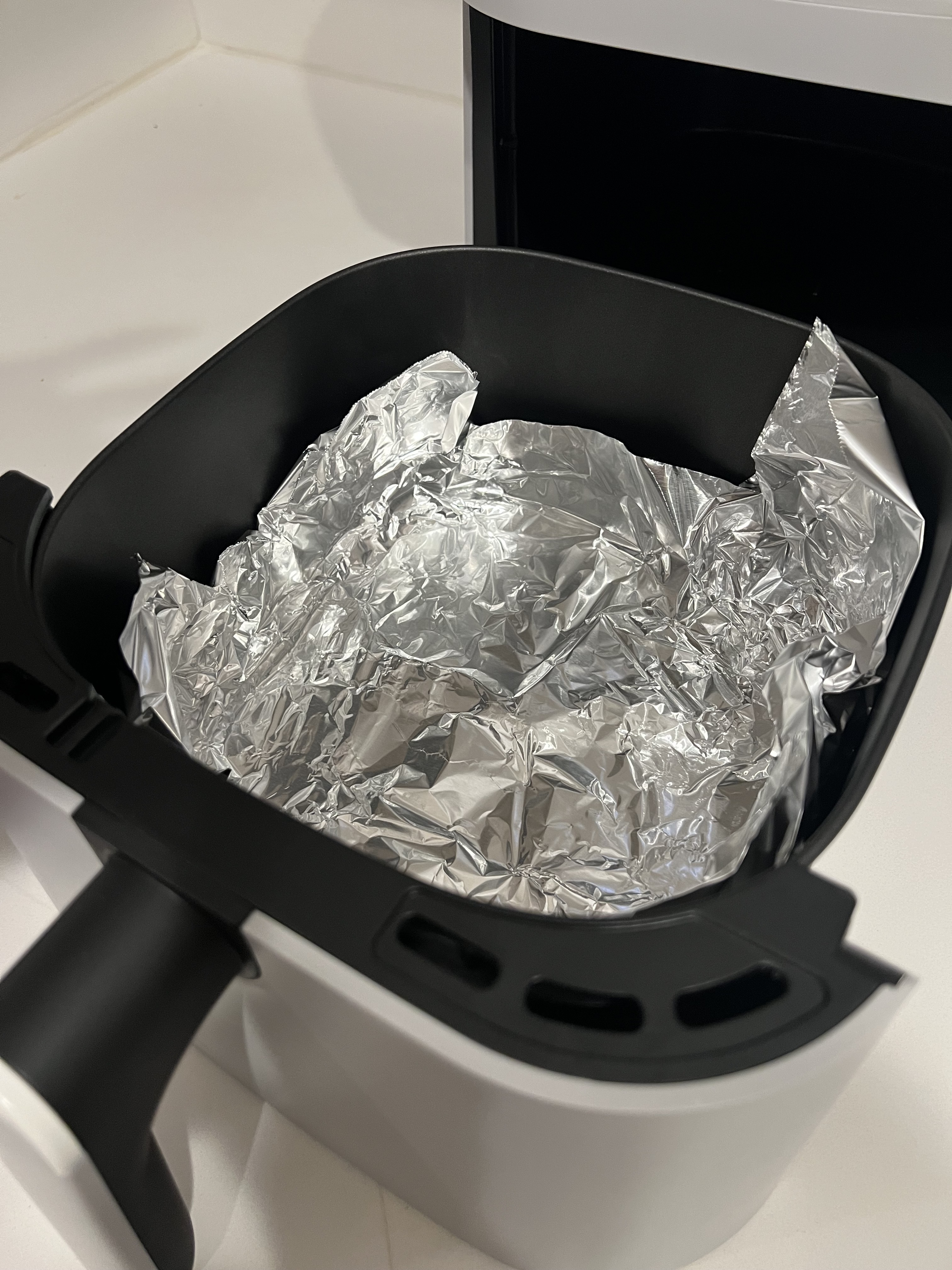 Air fryer basket lined with crumpled aluminum foil. The appliance is open, showing the interior