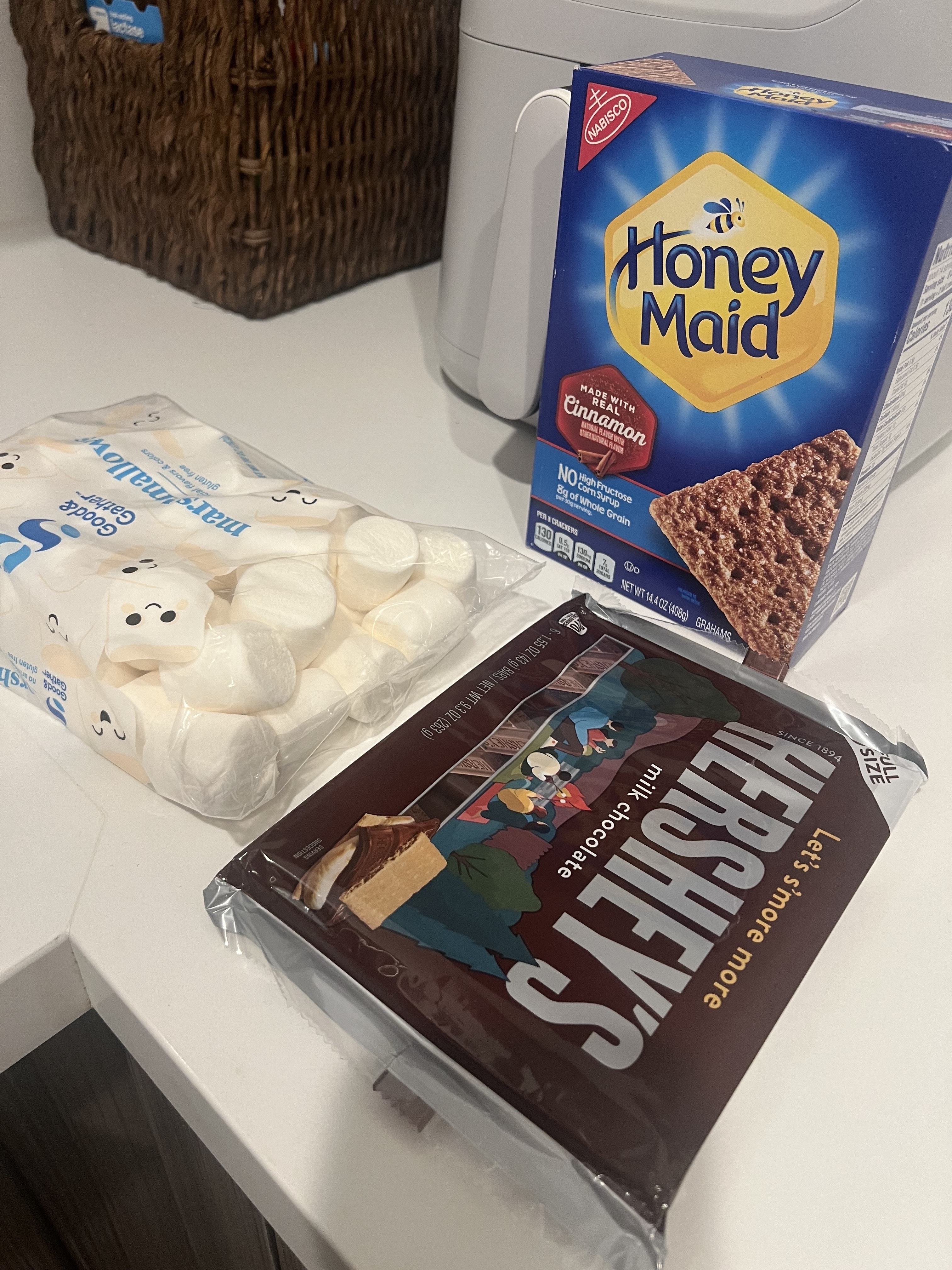 Packaged food items on a kitchen counter: Honey Maid cinnamon graham crackers, Jet-Puffed marshmallows, and Hershey's milk chocolate bars